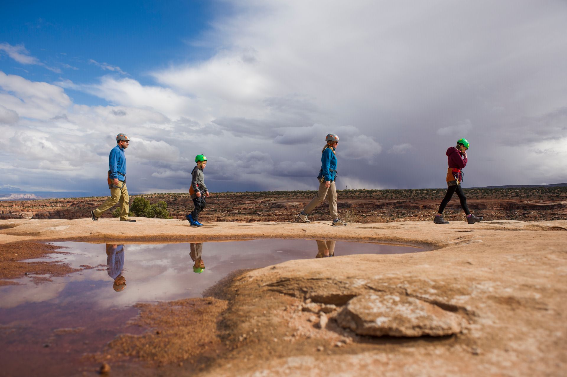 Four hikers with backpacks walk along a muddy desert trail under a cloudy sky.