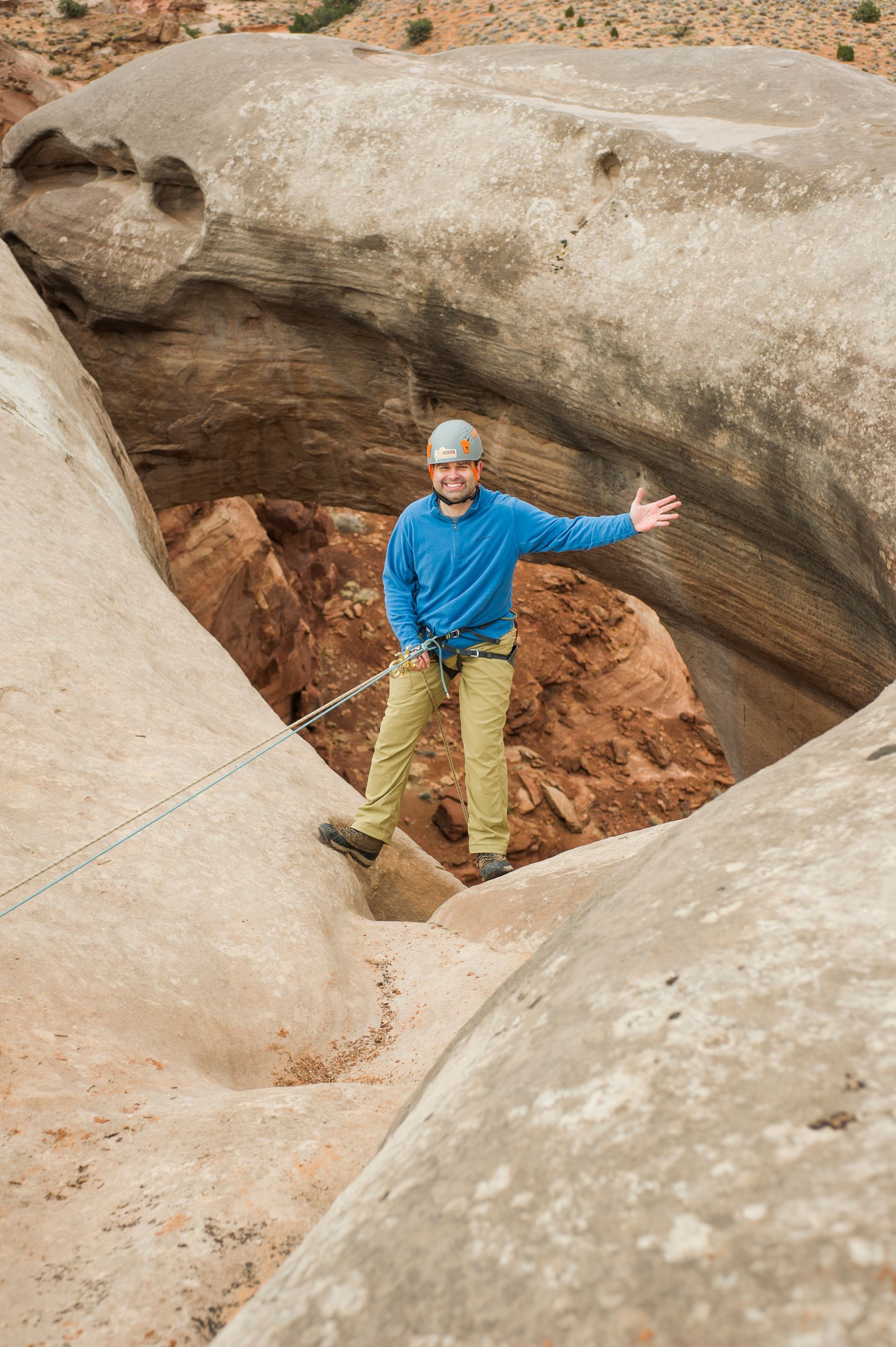 A smiling person wearing a helmet and blue jacket rappels through an arch rock formation while connected to a climbing rope on a guided All Ways Adventure tour in Kanab Utah near Zion.