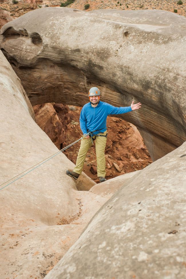 A smiling person wearing a helmet and blue jacket rappels through an arch rock formation while connected to a climbing rope on a guided All Ways Adventure tour in Kanab Utah near Zion.