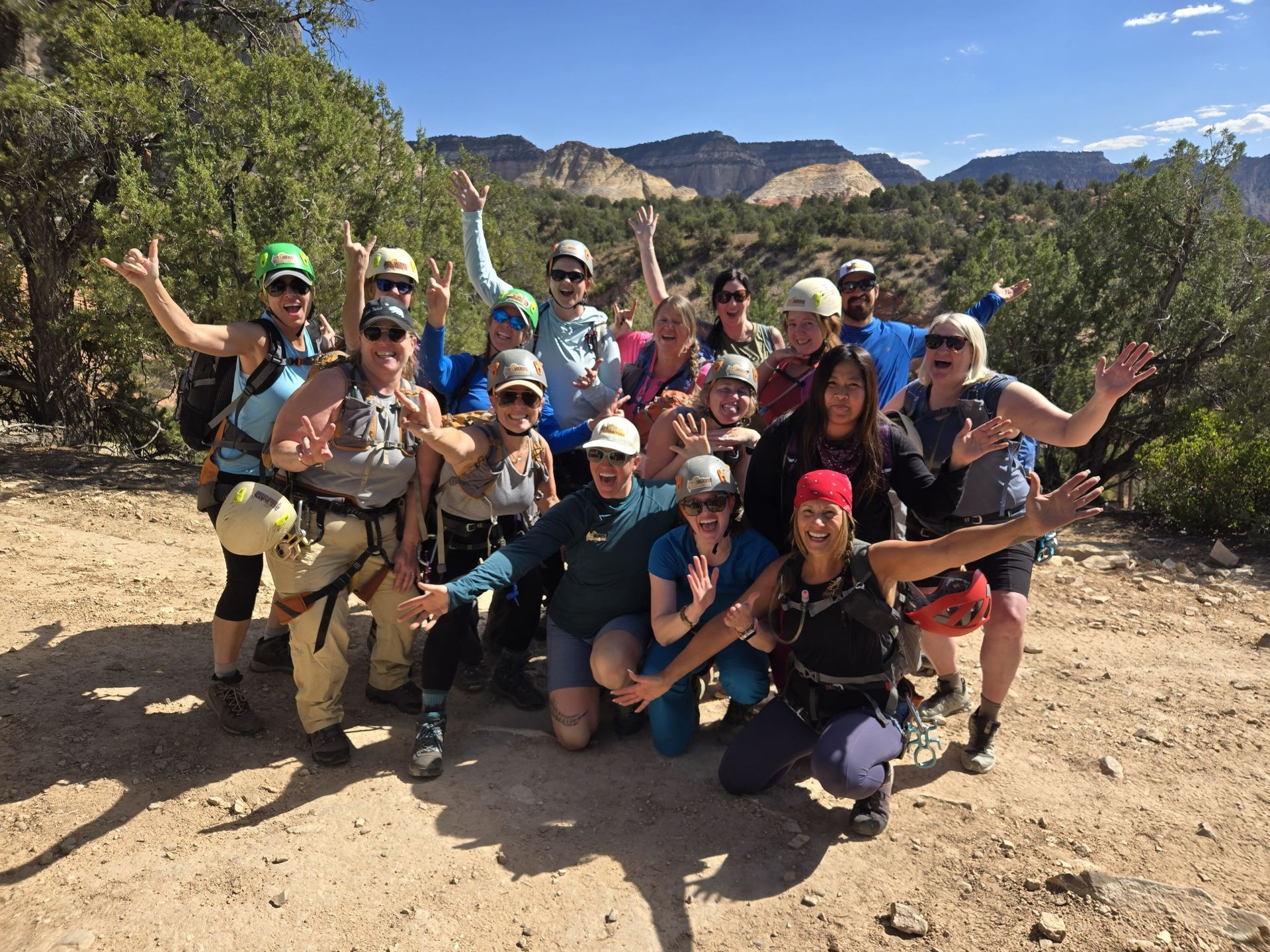 A group of hikers wearing helmets and climbing harnesses poses outdoors on a dirt path with rocky hills in the background.