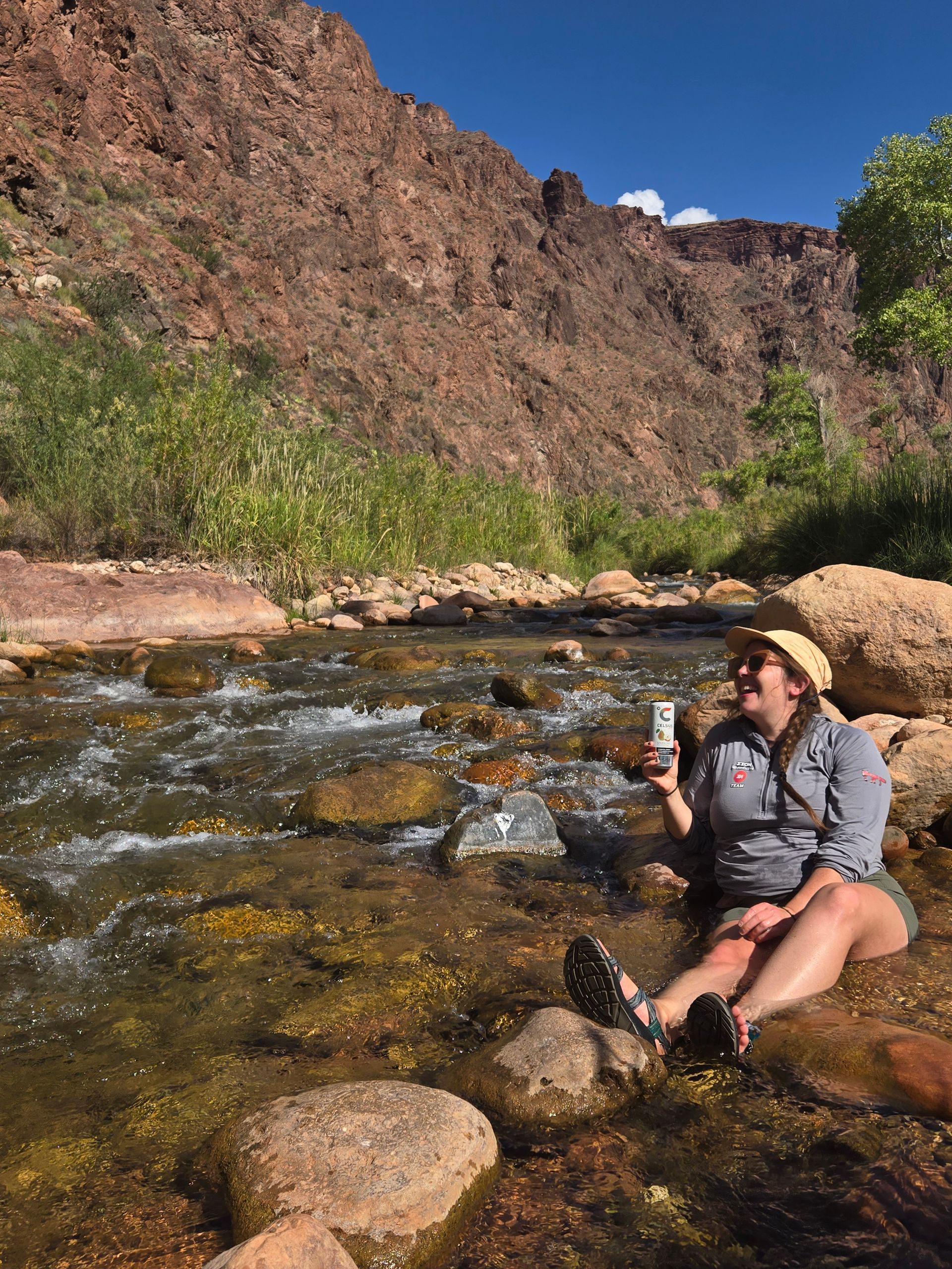A person sits in a stream with a cool drink