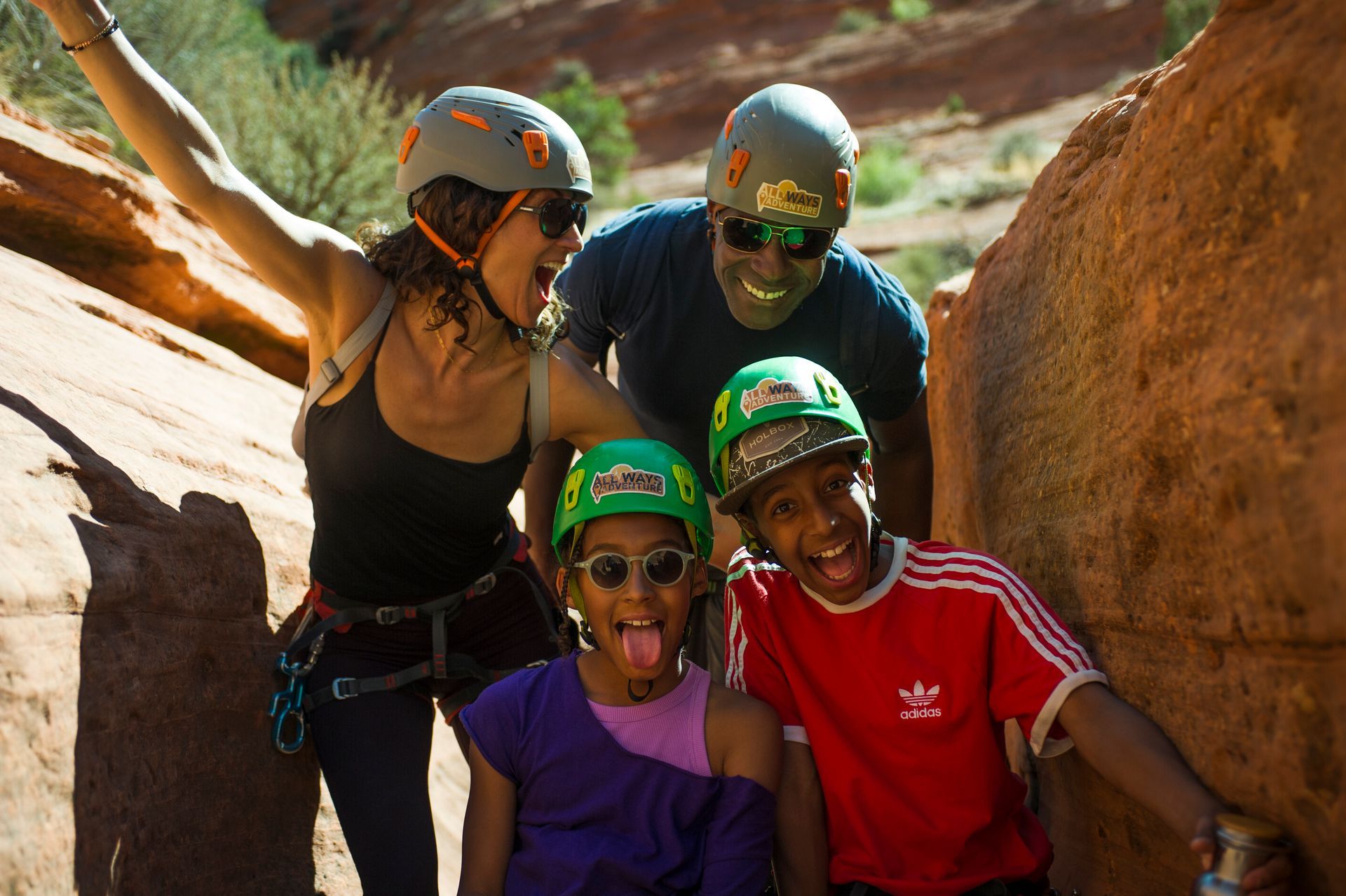 A family wears helmets and climbing gear, smiling excitedly while hiking between red rock canyon walls.