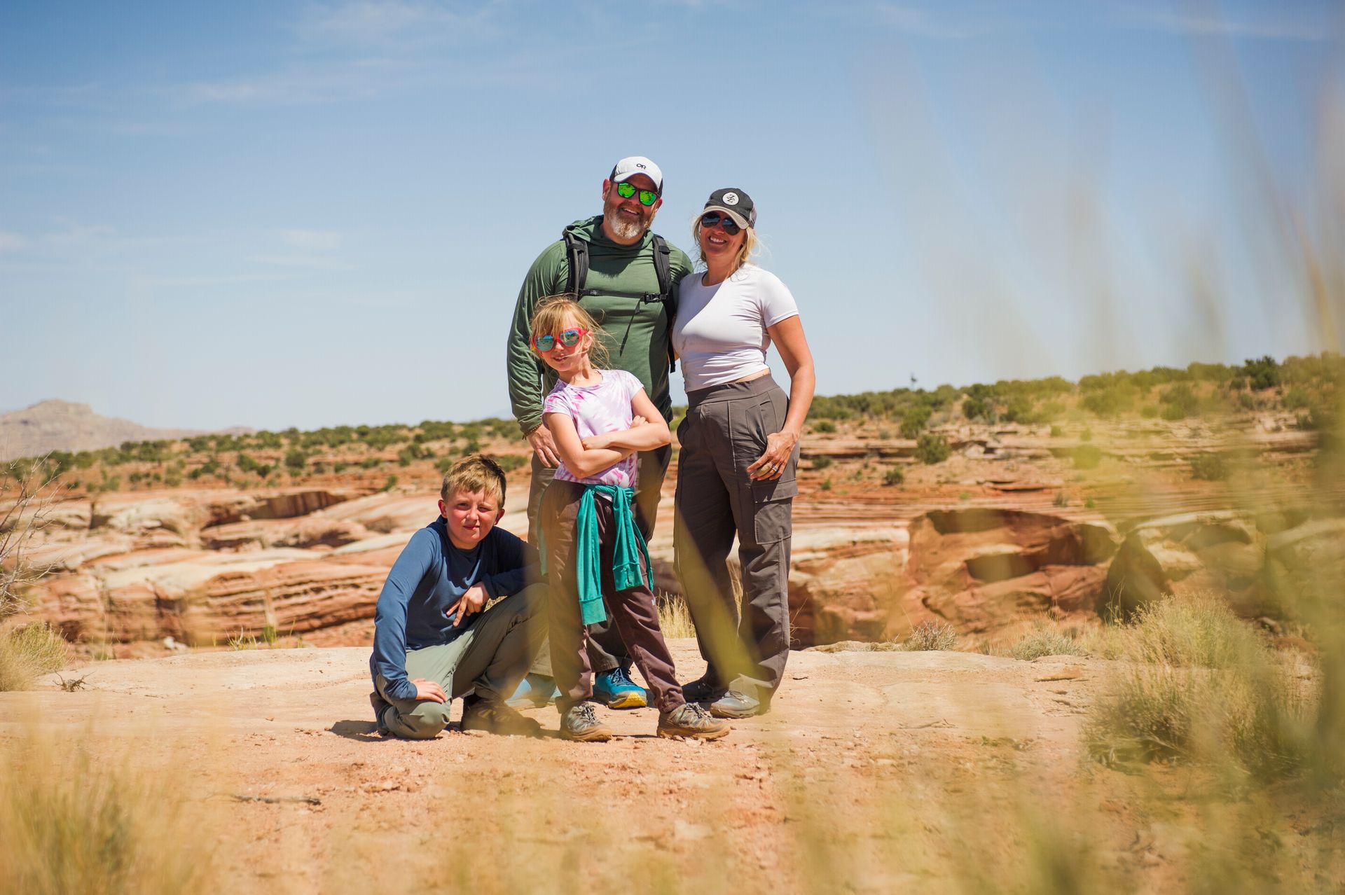 A family of four poses together on a sunny desert trail with rocky cliffs in the background.