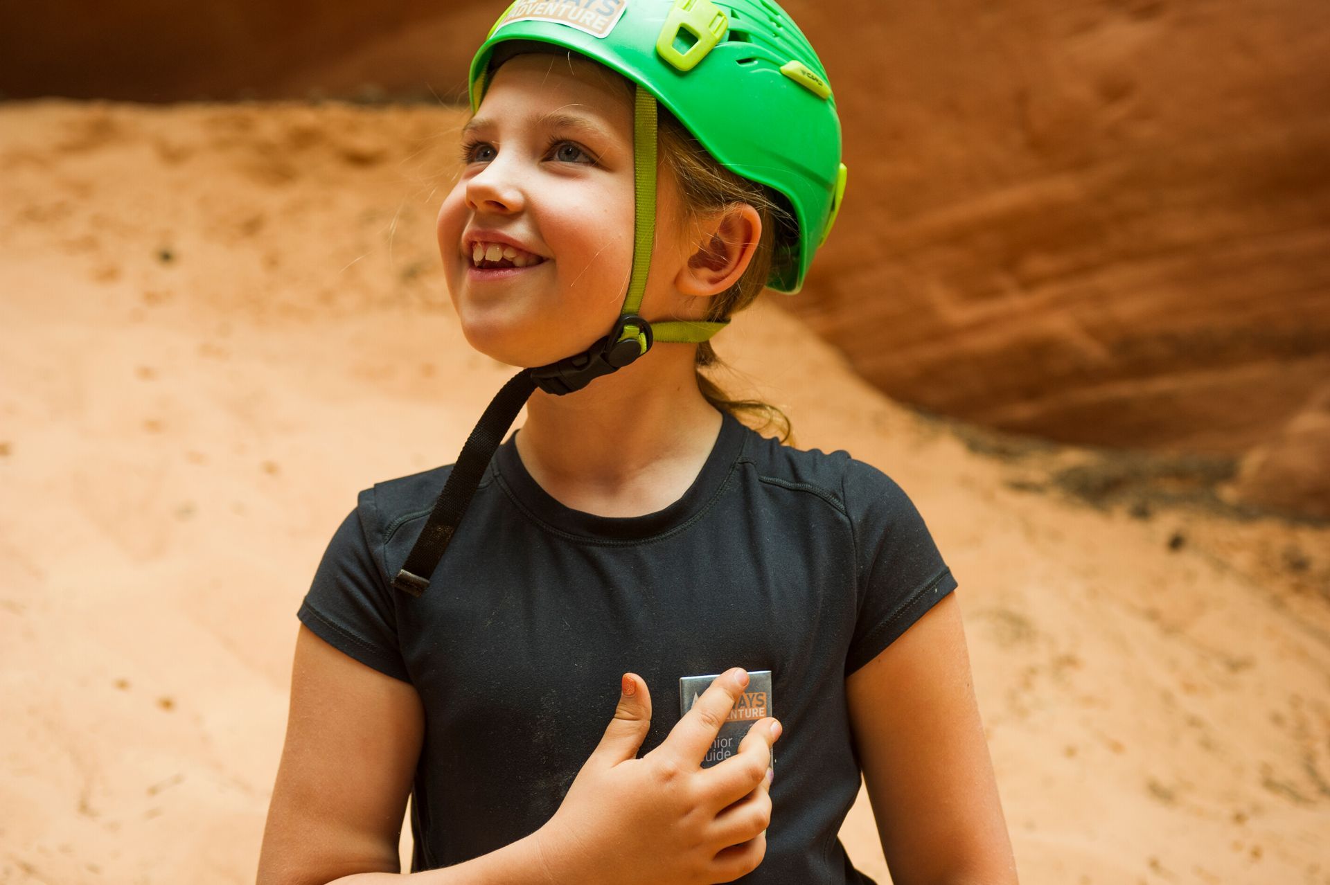 A smiling person wearing a green climbing helmet stands in a sandy, rocky canyon.