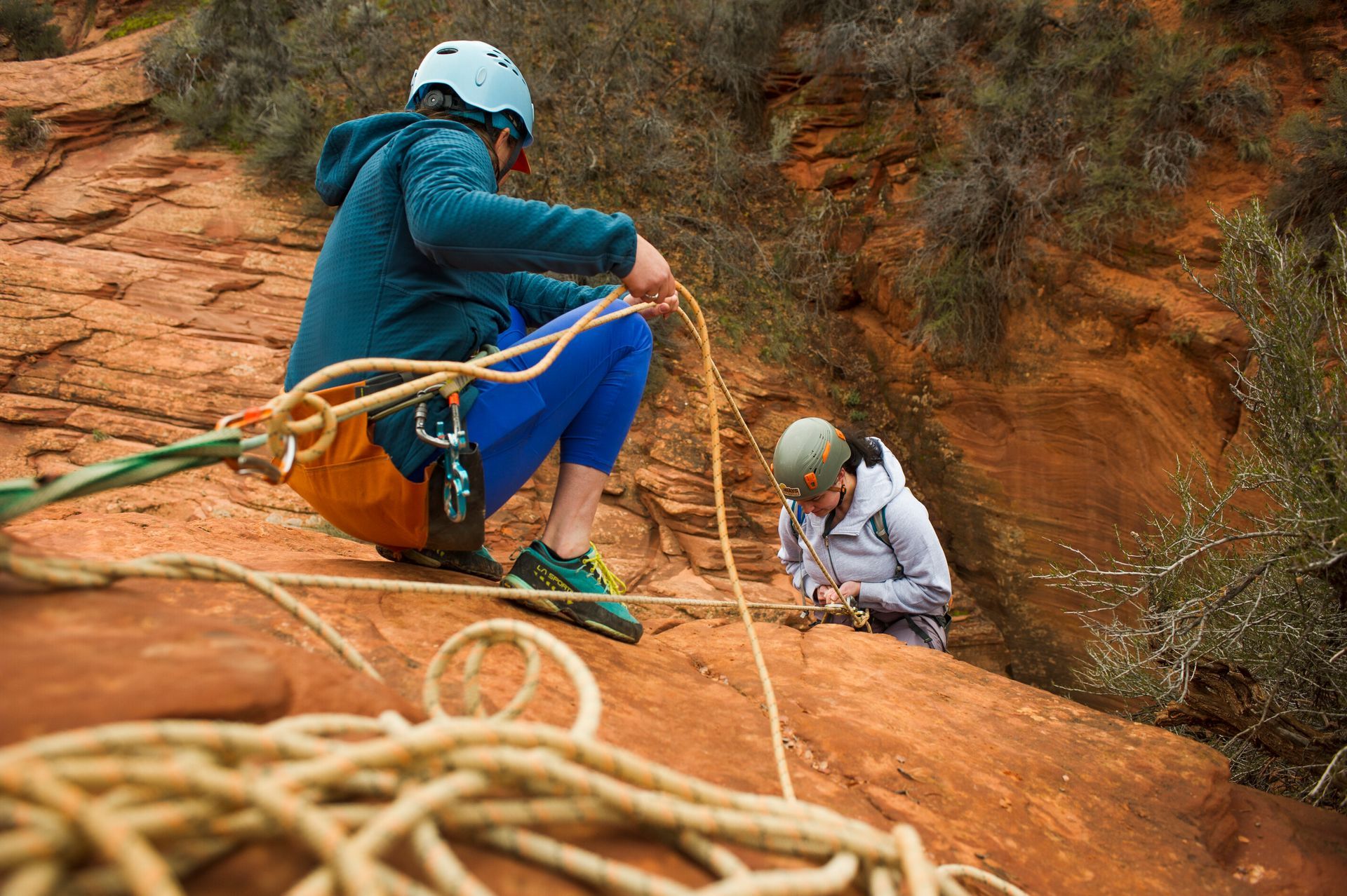 Two people preparing for a climb on a red rock formation, one handling rope, the other adjusting gear.