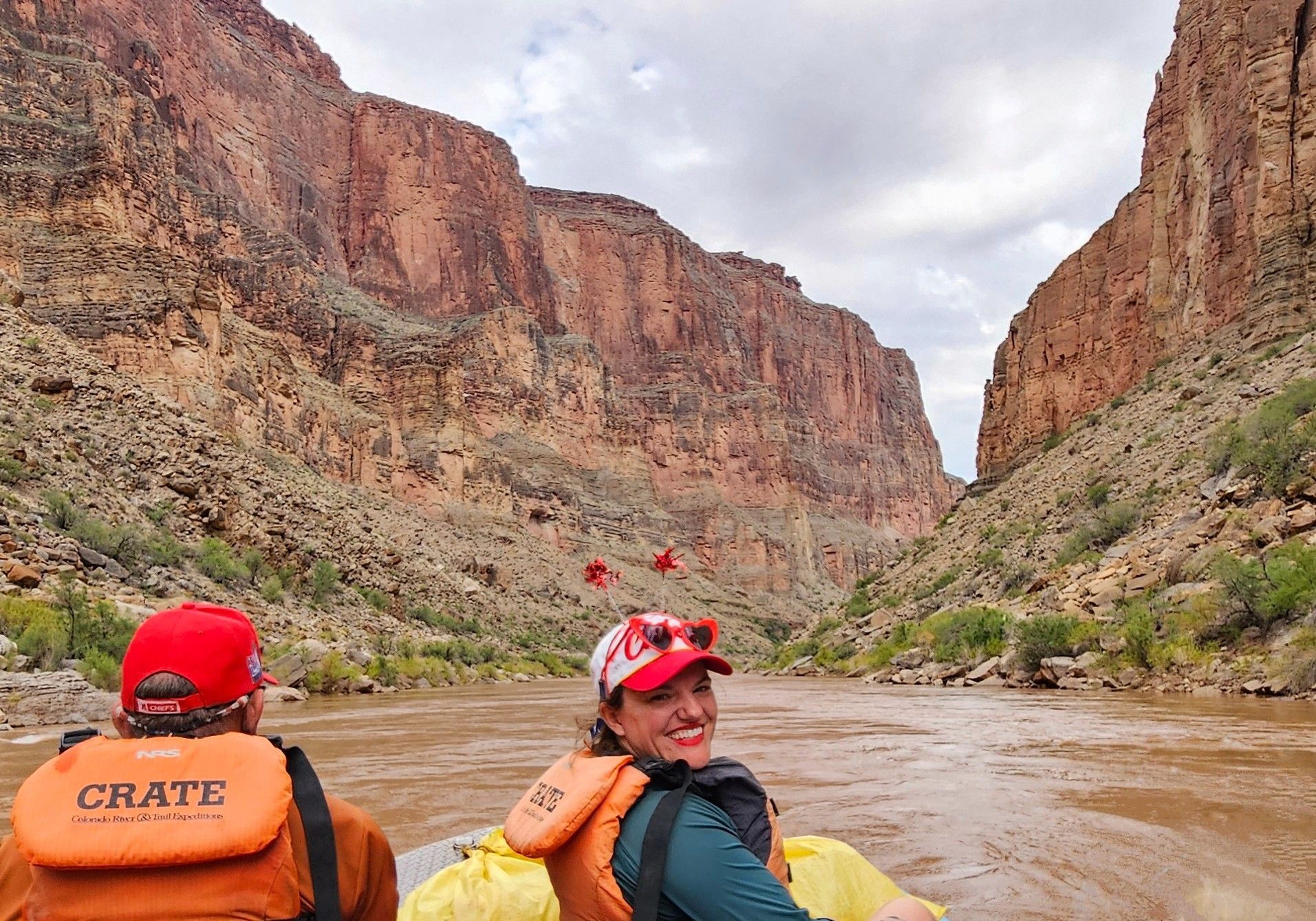 People in raft on river, smiling. Red-rock canyon walls rise on either side. Cloudy sky.