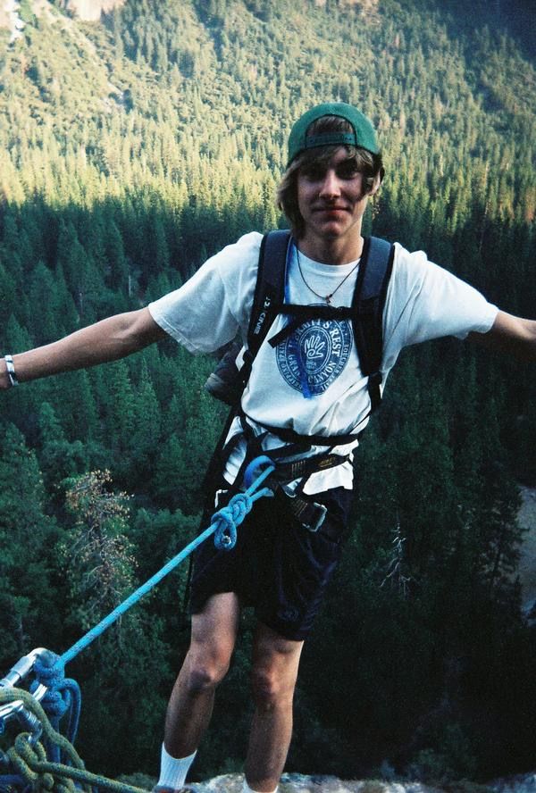 Nathan wearing a green hat, a backpack, and a harness is tied to a rope and leaning over a forest. 