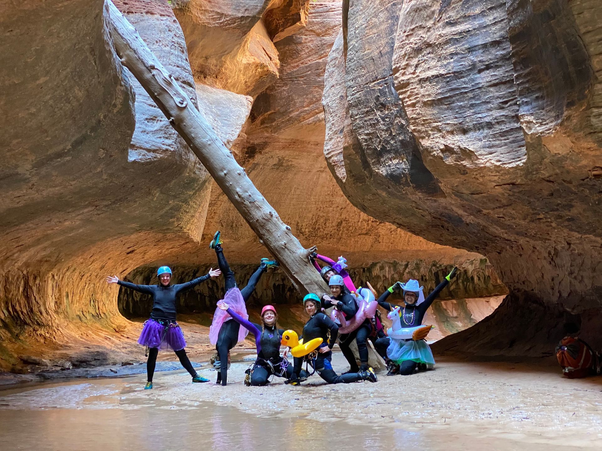 A group wearing colorful tutus and a duck floatie poses under a large log spanning a narrow, sandstone slot canyon.