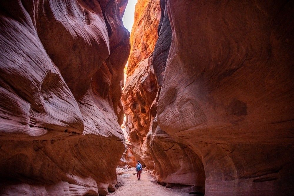 A person hikes through a narrow, sandstone canyon, illuminated by sunlight from above.