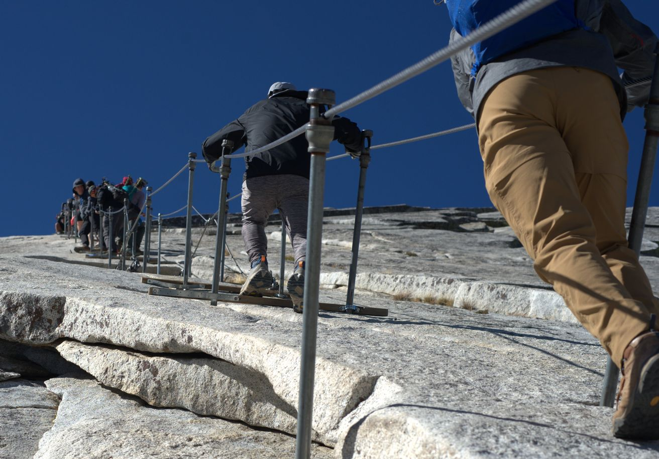 People climbing steep granite with cable handrails under a clear blue sky.