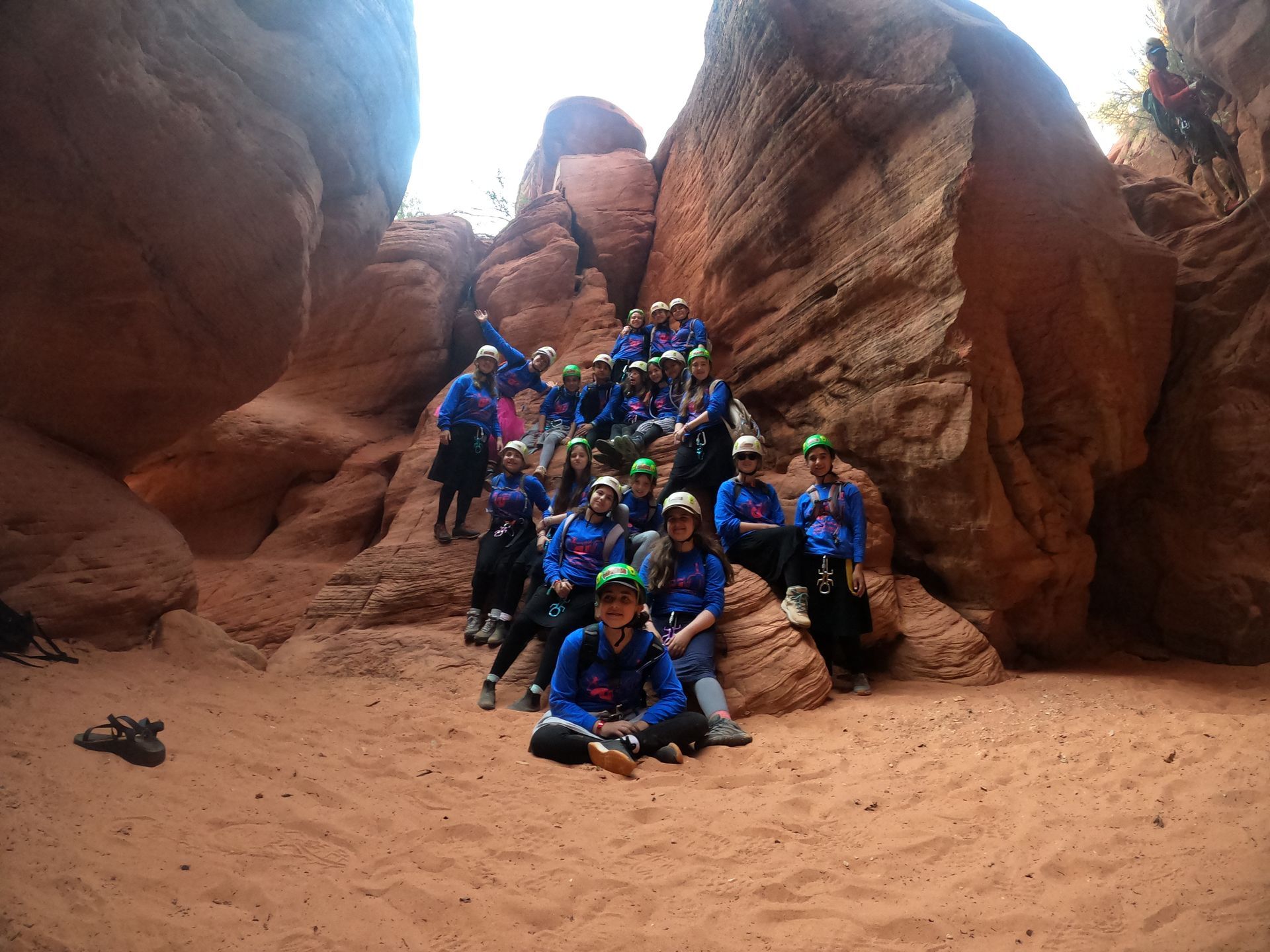 A group of people in blue shirts and hats pose in a red rock canyon. Some are sitting, others standing, smiling.