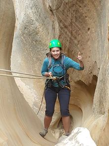 Woman rappelling in a sandstone canyon, wearing a green helmet, blue shirt, and giving a thumbs-up.