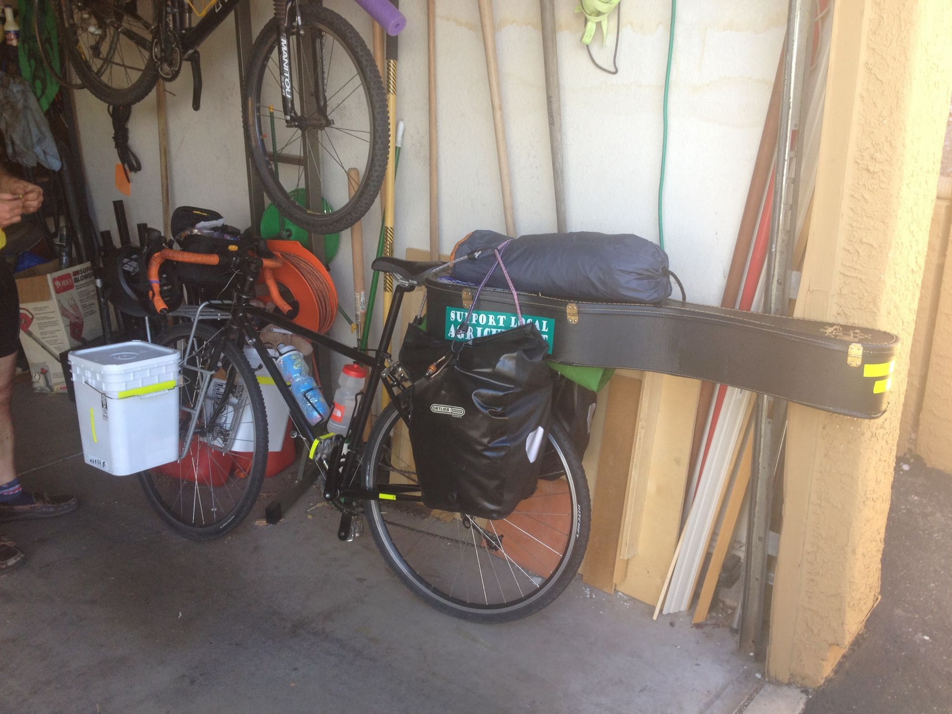 A black bicycle loaded with gear, parked inside a garage. A board and other items lean against the wall.
