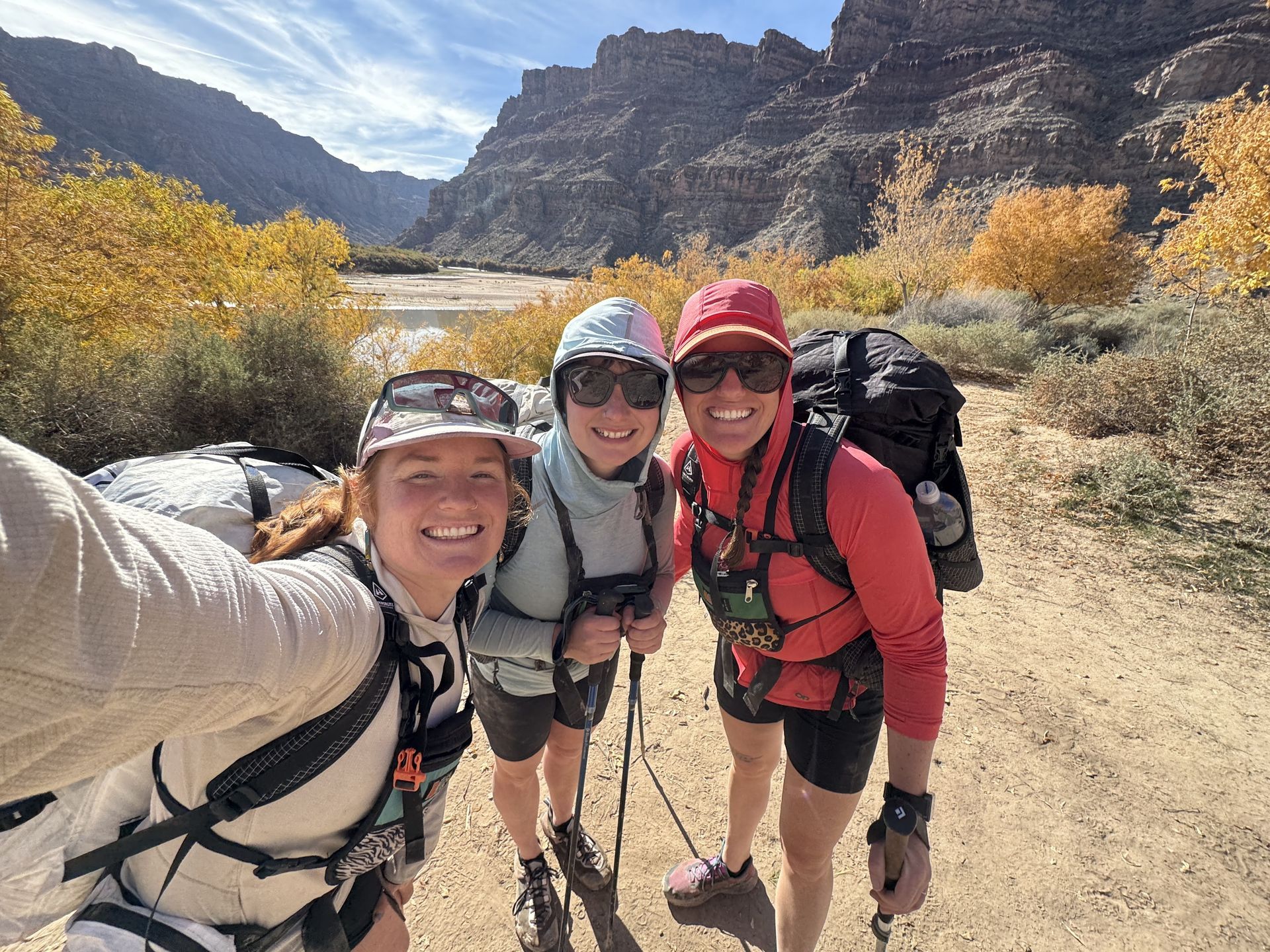 Three hikers on a sunny trail pose for a selfie. Yellow trees and canyon backdrop.