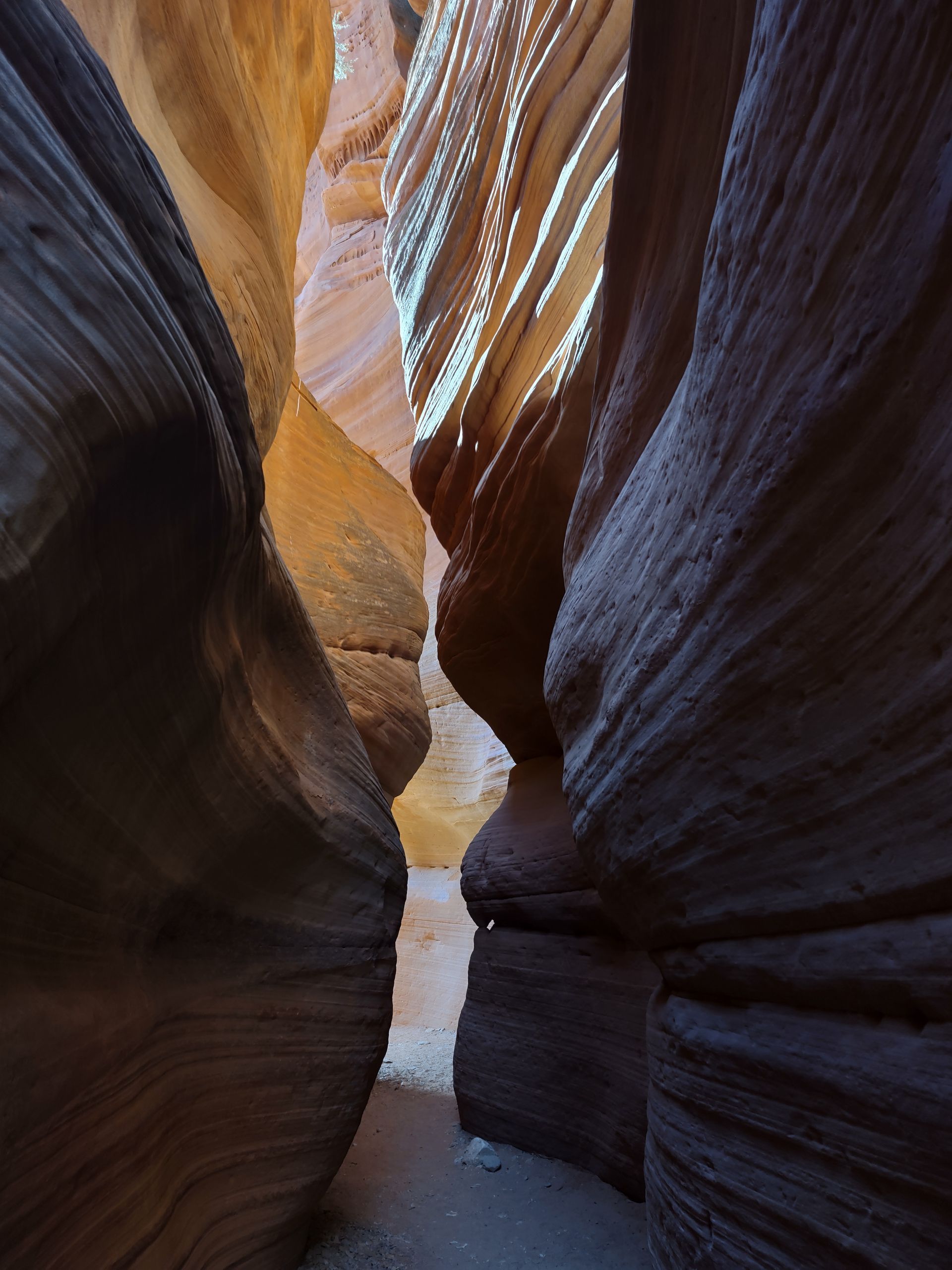 A narrow slot canyon with light-colored walls and a sandy floor, lit from above.