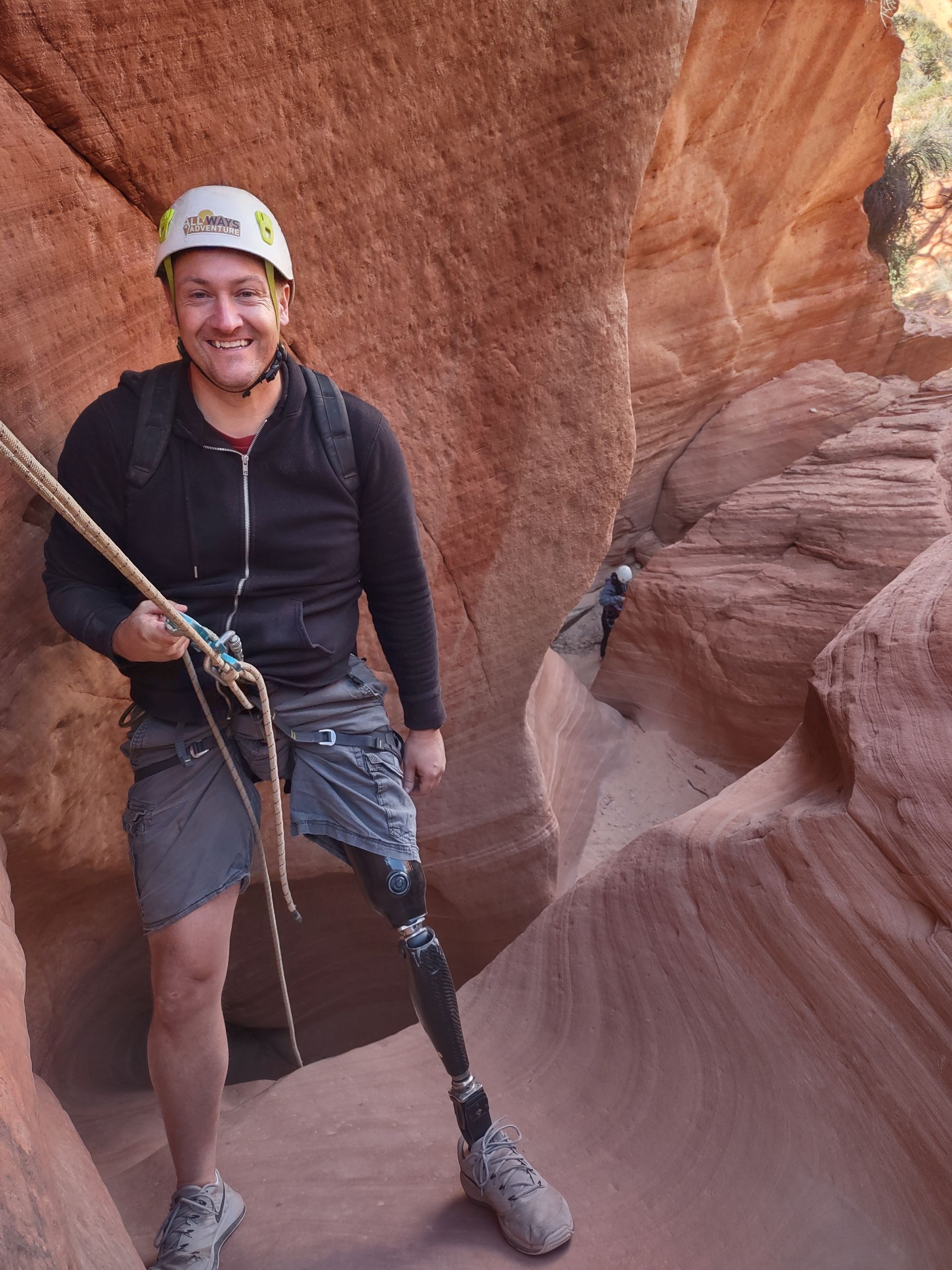 A man with a prosthetic leg smiles, holding a rope in a red rock canyon. He wears a helmet, backpack, and climbing gear.