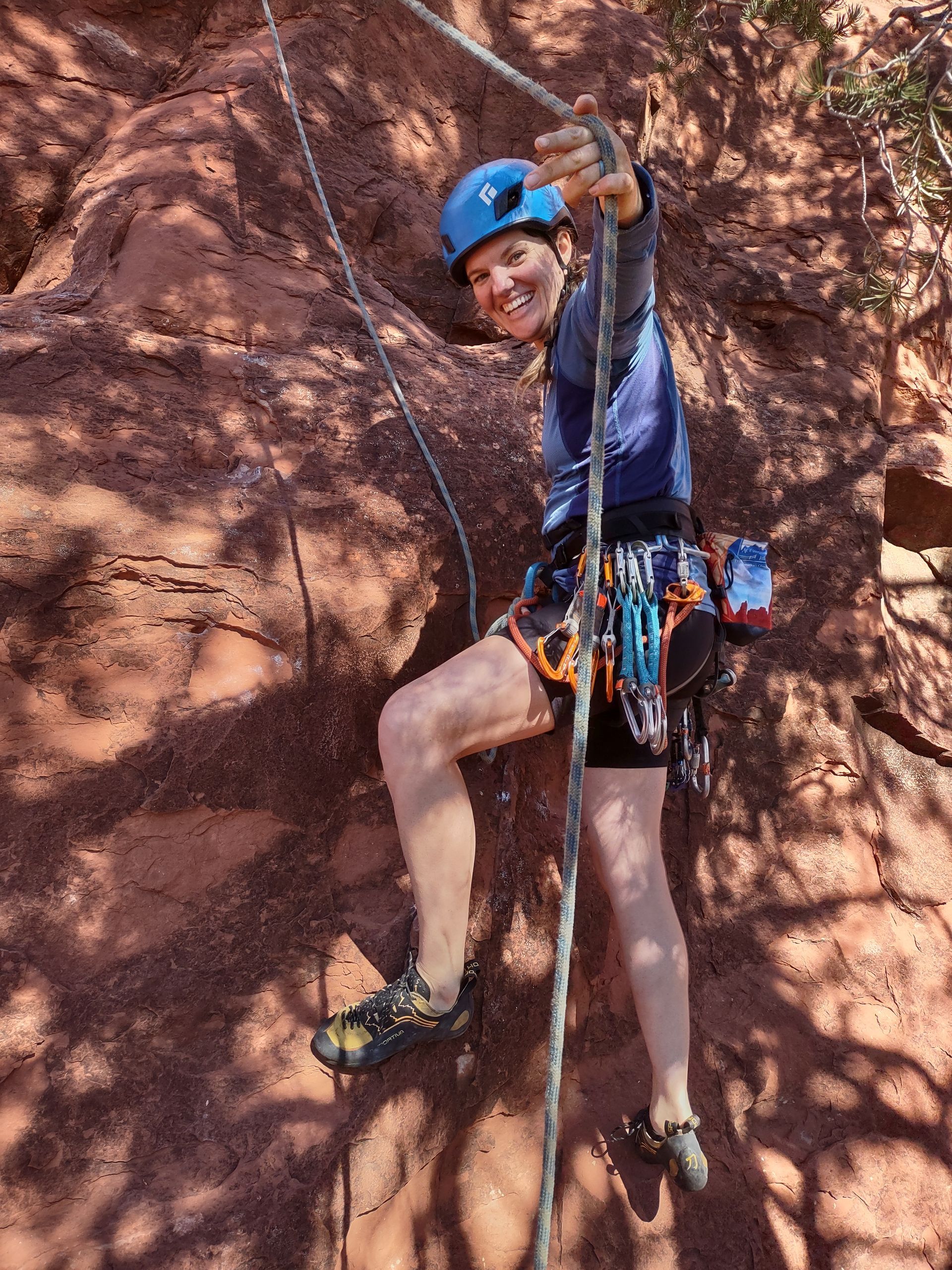 A person is smiling and holding a rope while standing on a red rock wall. 