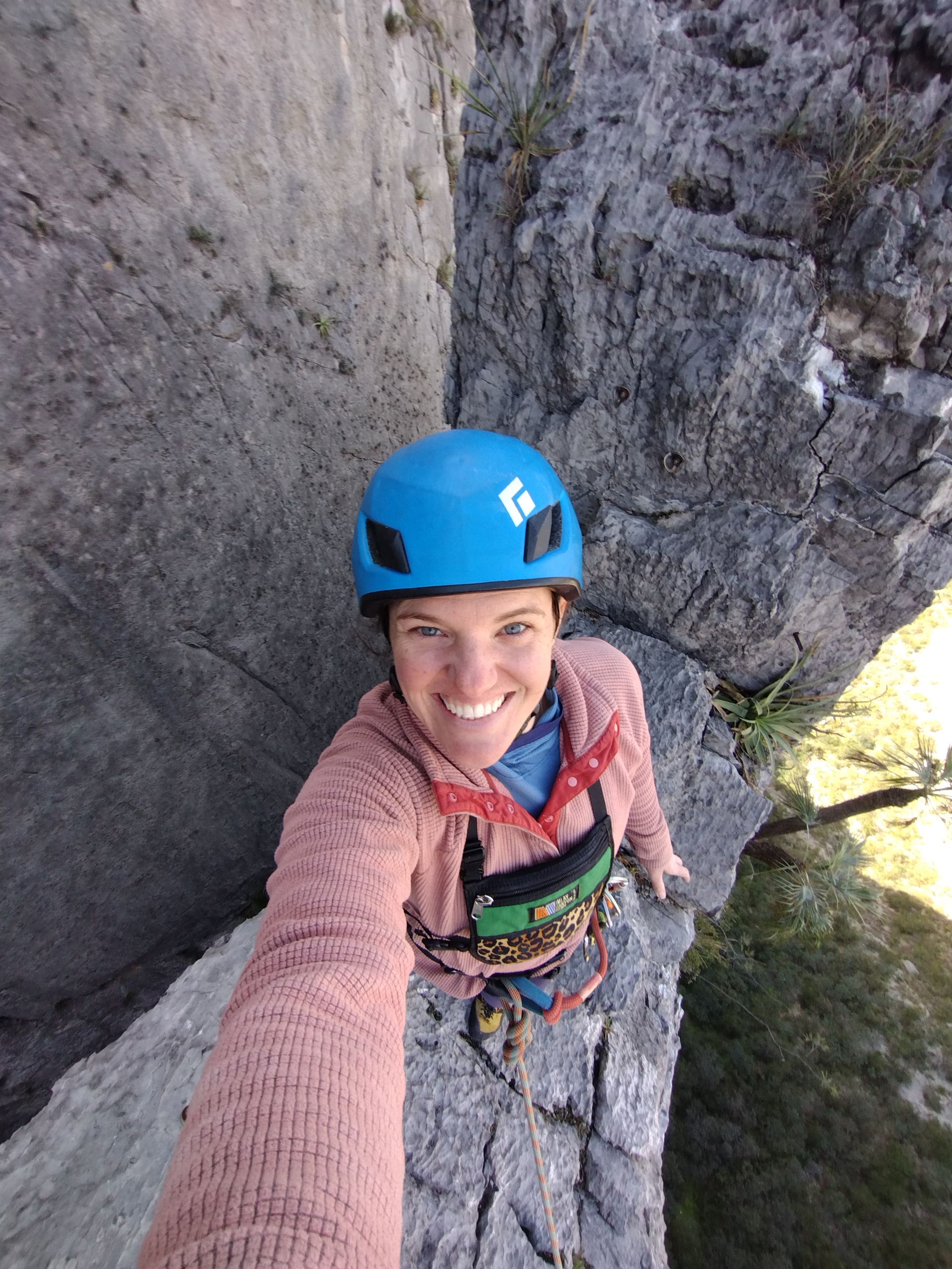 Jillian is wearing a blue helmet and taking a selfie while standing on a ridgeline with cliffs on both sides behind her