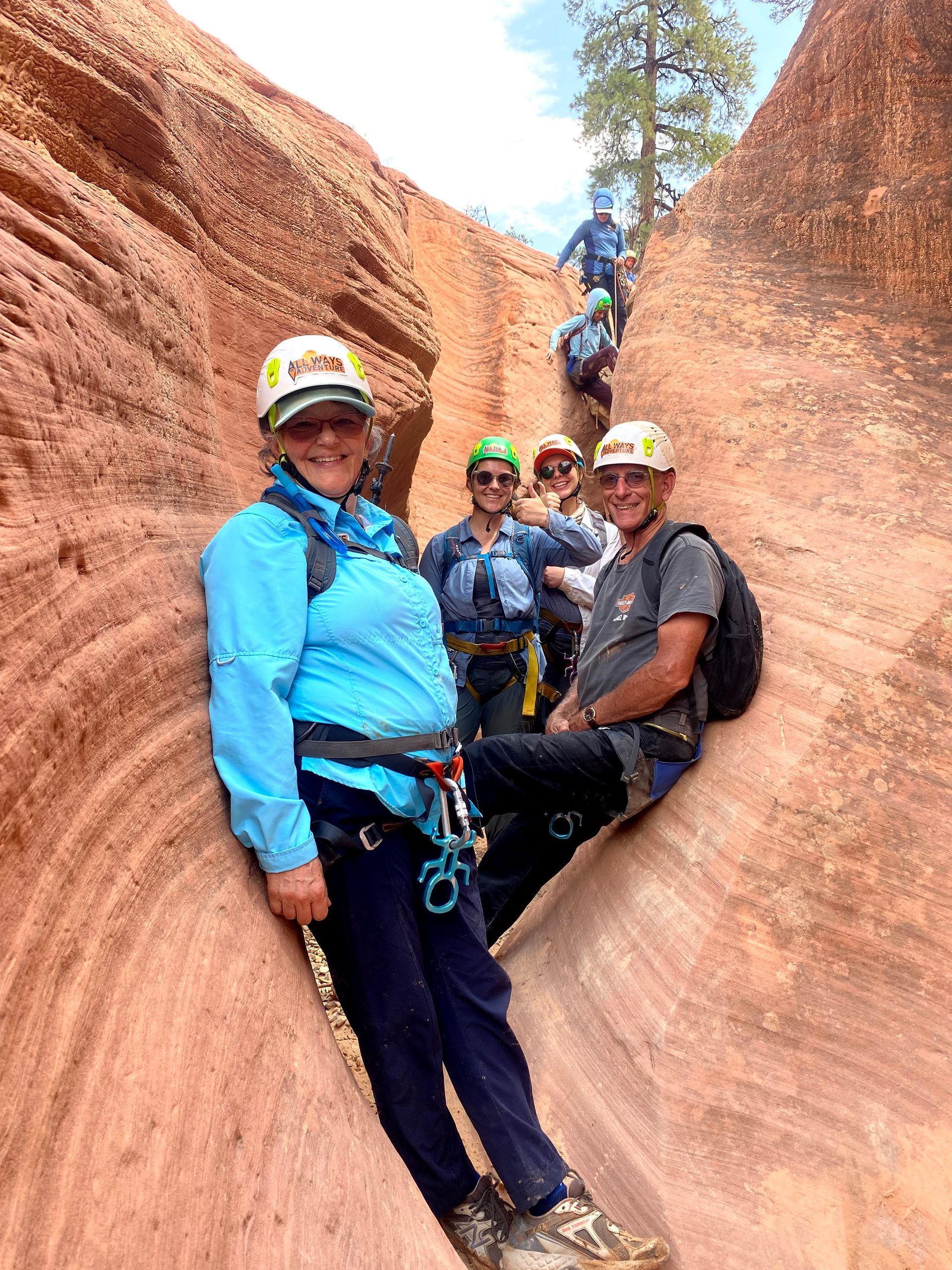 Group of people canyoneering in a narrow red rock canyon. Two in foreground smiling, wearing helmets and harnesses. Others climbing up.