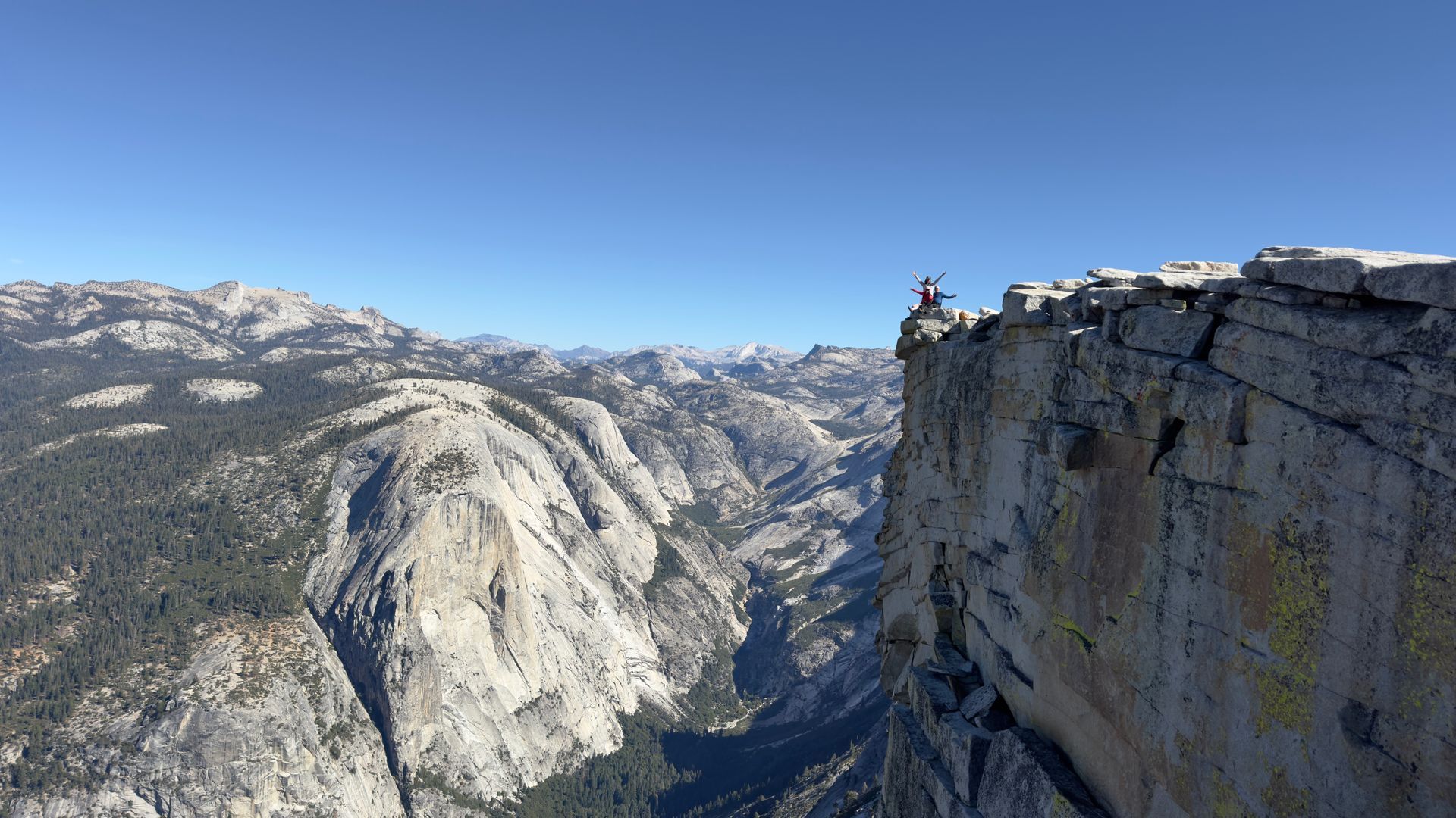 Person standing on the edge of a cliff, overlooking a valley. Sunny, blue sky.