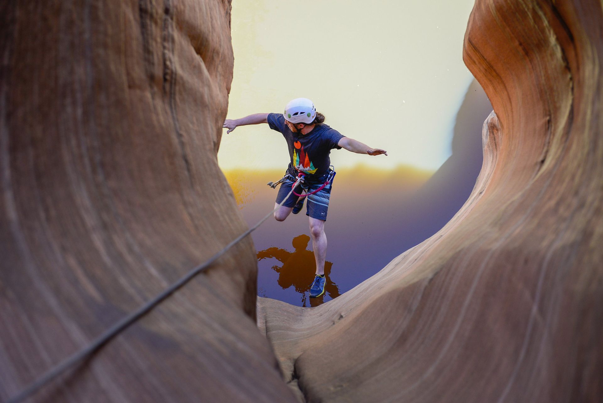 Man balancing on a rope bridge in a desert canyon, arms outstretched, wearing a helmet and harness.