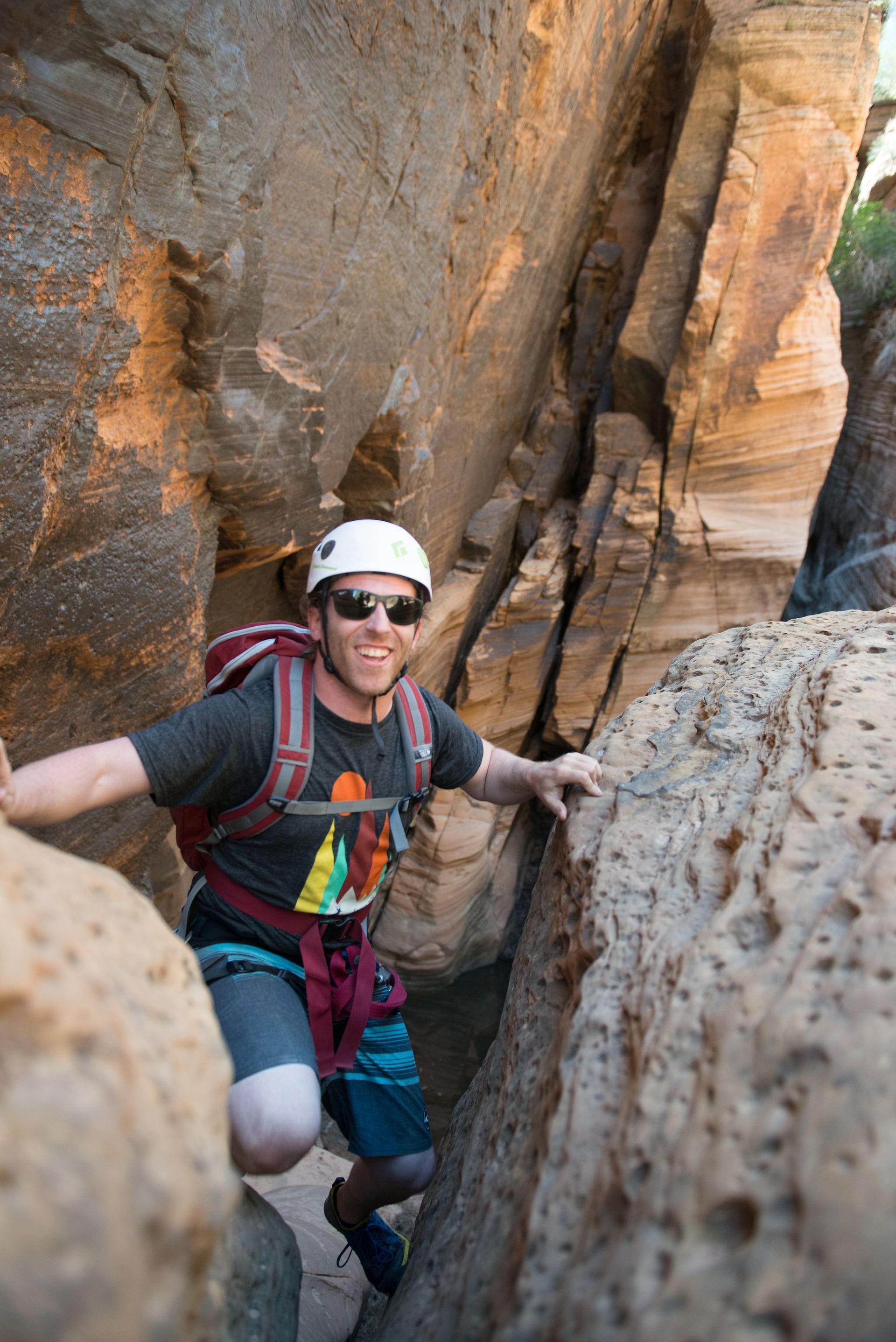 Man in helmet and sunglasses smiles while climbing through a narrow rock canyon.