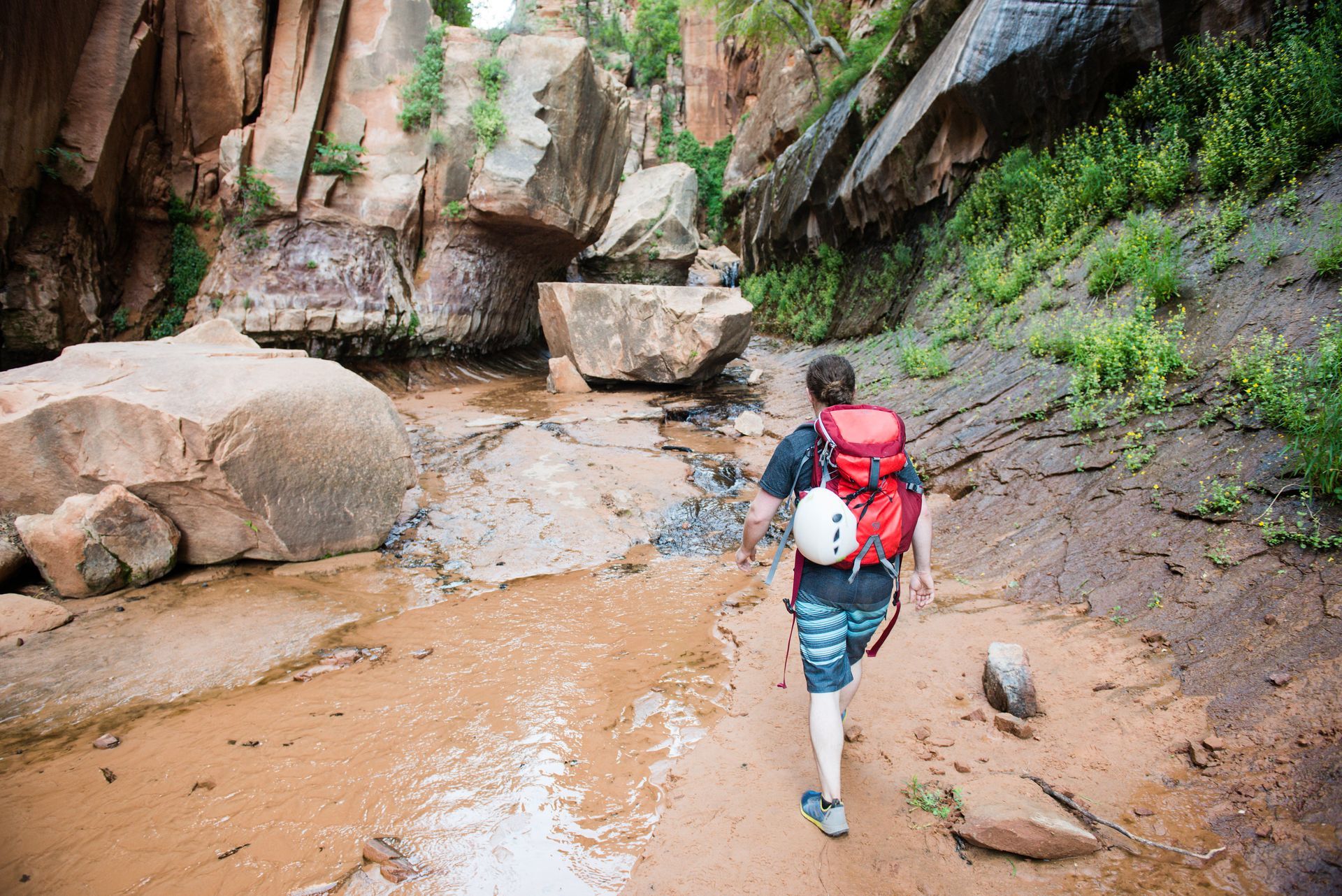 Person with red backpack hikes through a narrow canyon with large rocks.