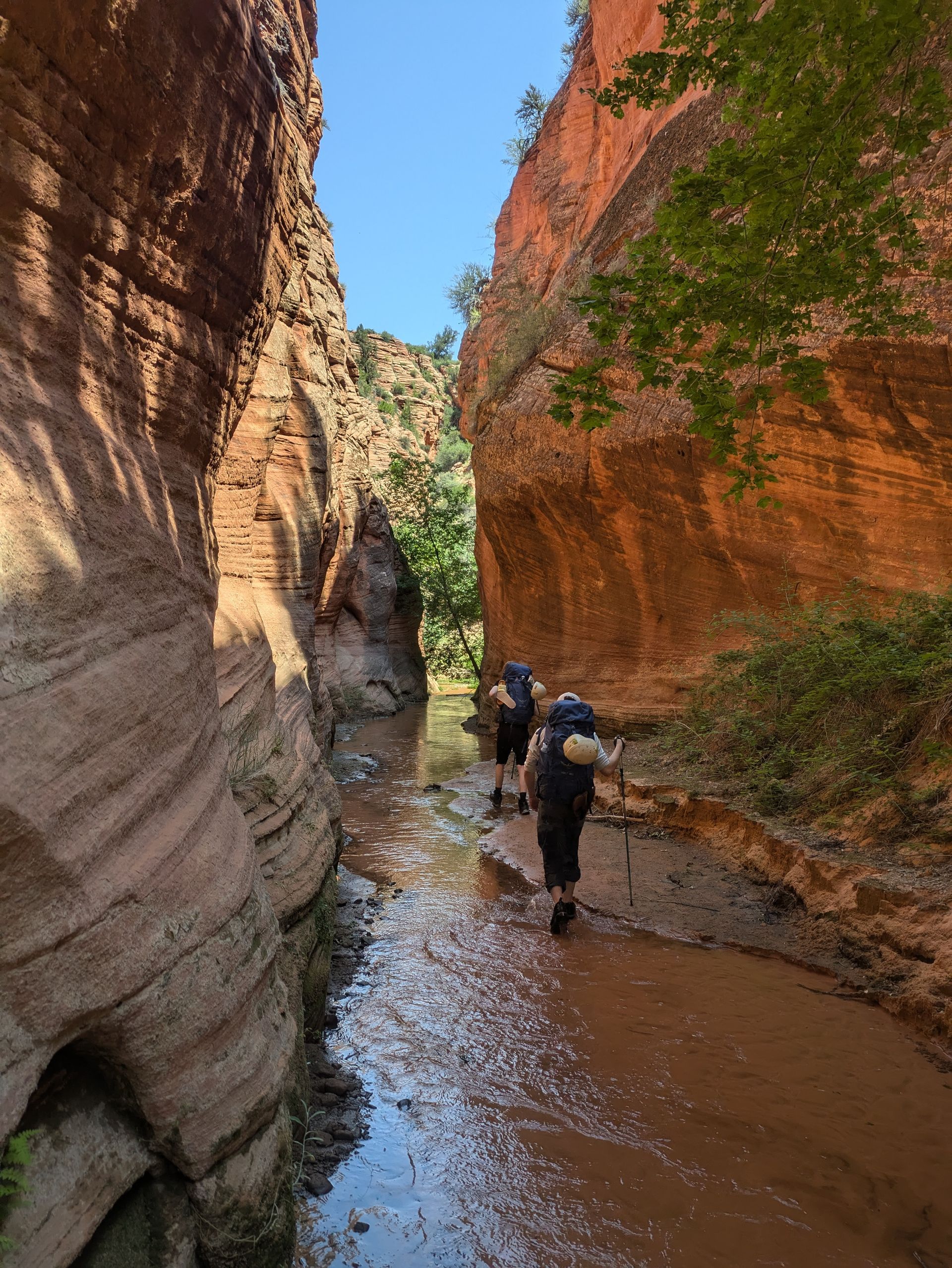 Two people with backpacks are walking through a muddy river