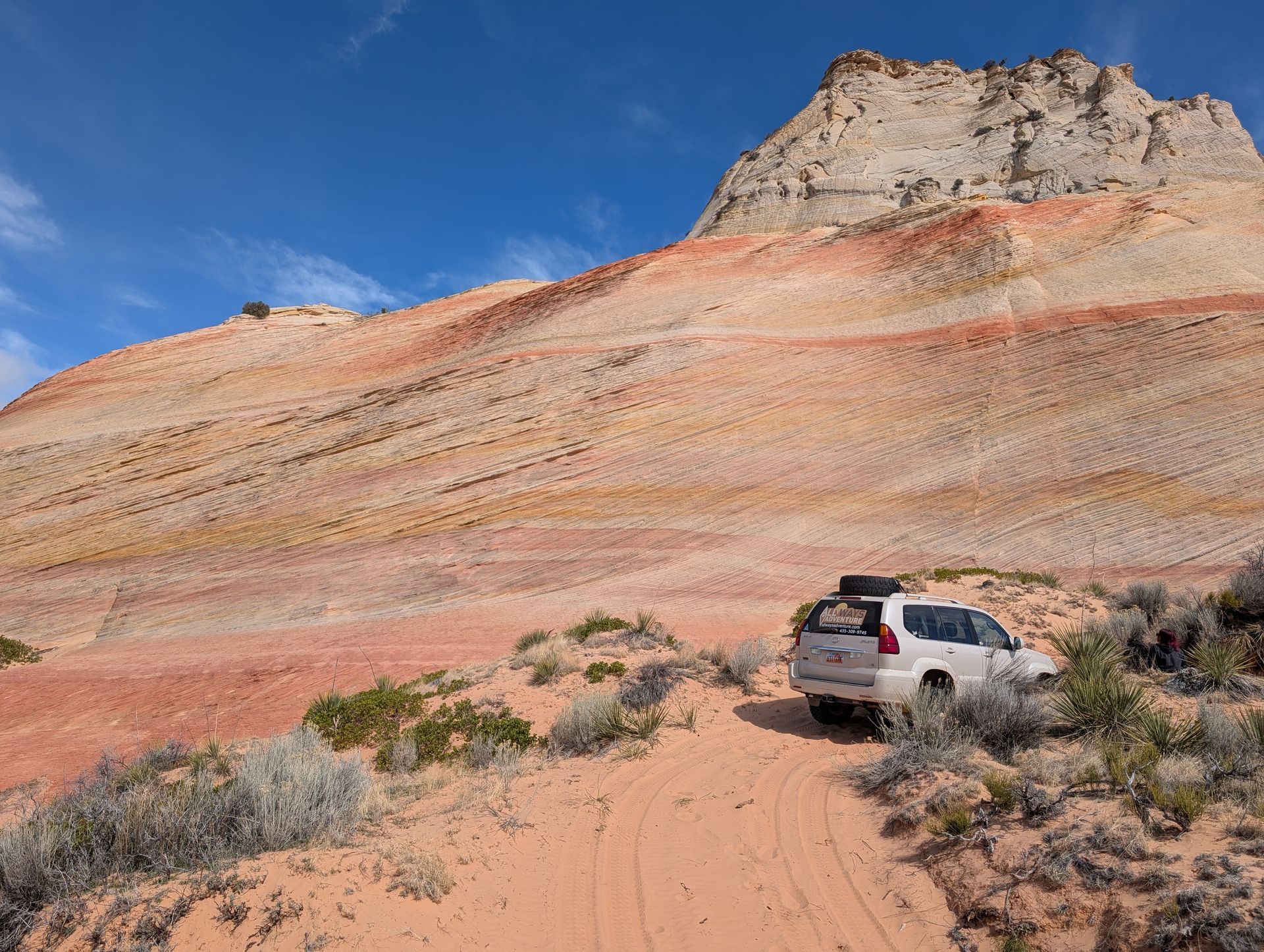 Tan SUV driving on a sandy path near a colorful, layered mountain under a blue sky.