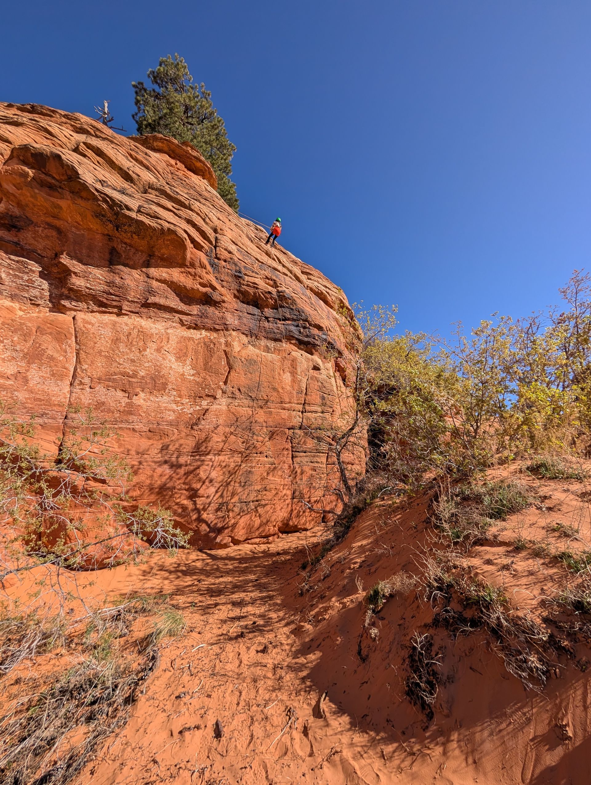 A person in a red shirt stands on a rocky, orange cliff ledge against a clear blue sky.