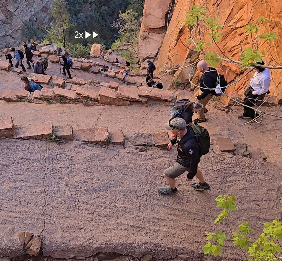 Hikers traverse a steep, winding path carved into a rocky cliffside in a canyon setting.