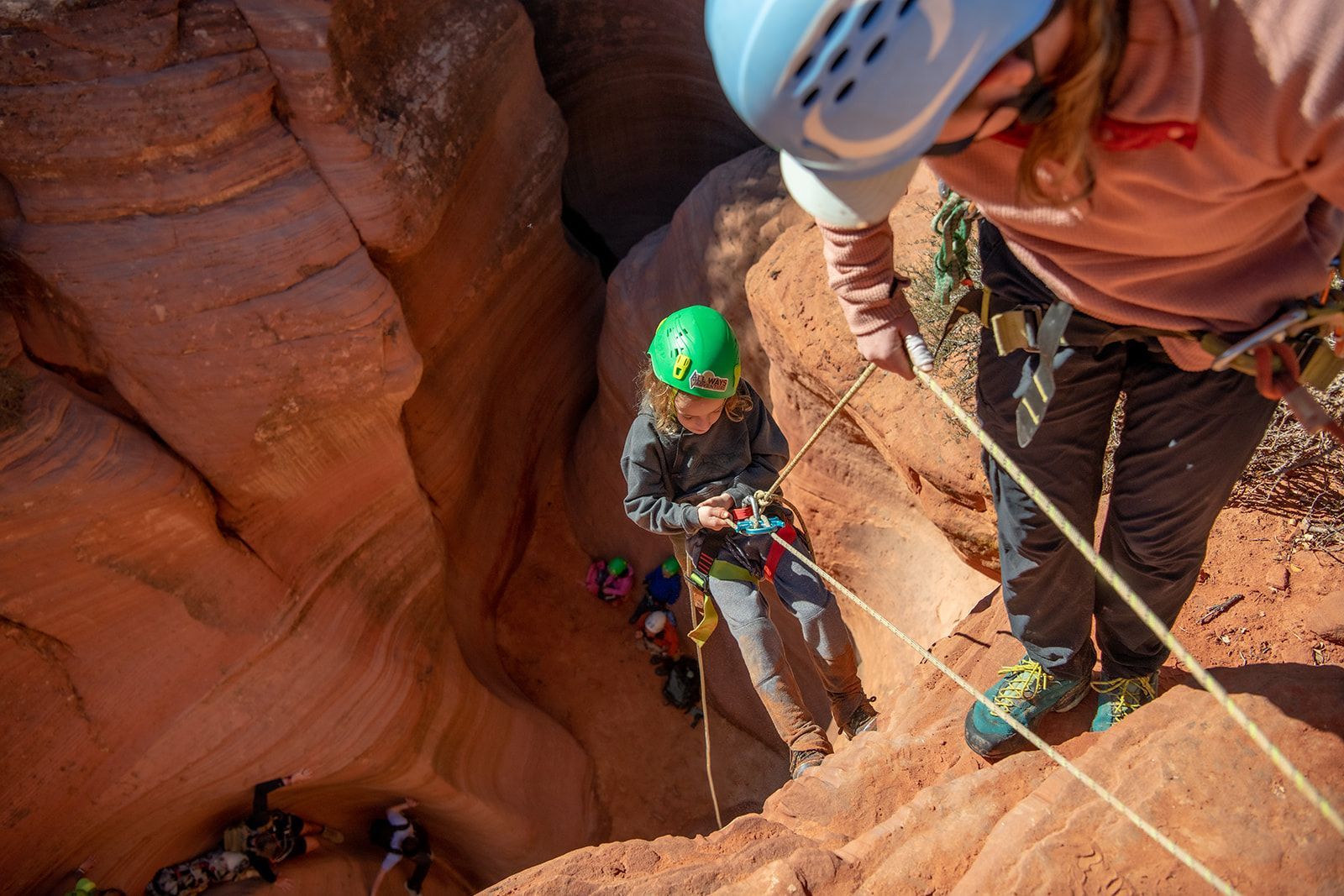 Two people rappelling down a red rock canyon. One wears a blue helmet, the other, green. Sunlight fills the canyon.