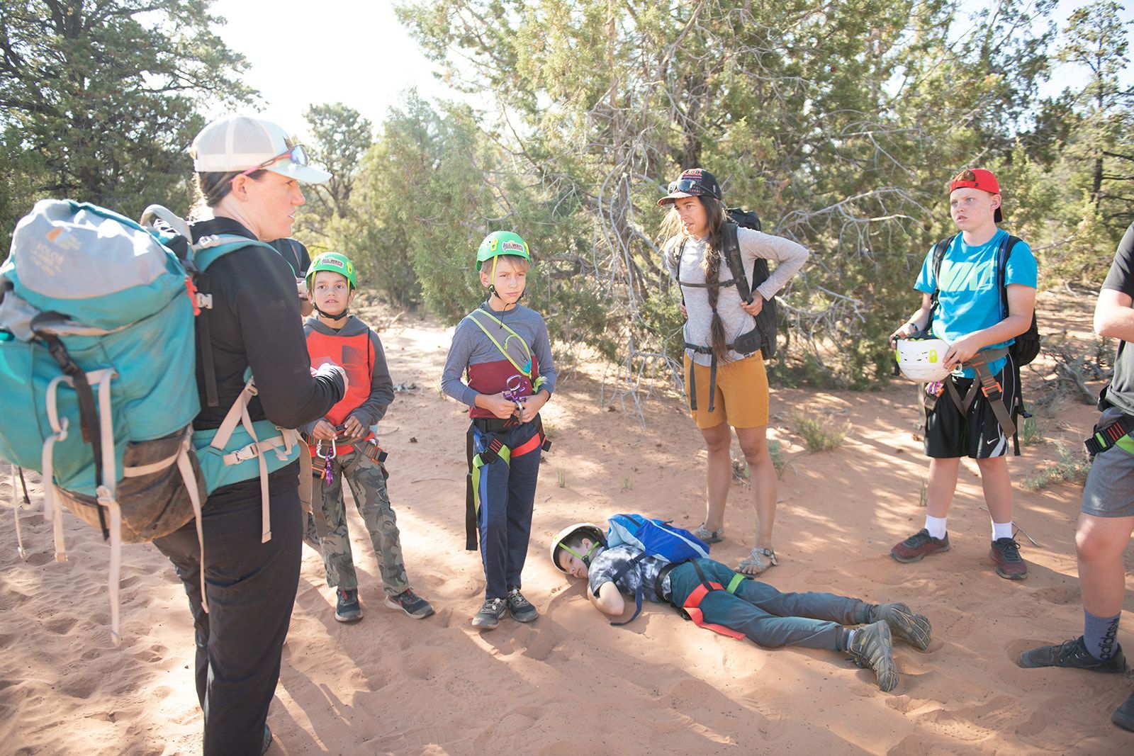 Group of people in climbing gear in a sandy outdoor area. One person lies on the ground, while others look on.