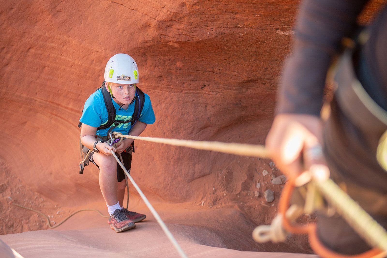 Boy rappelling down a red rock canyon, holding a rope with a harness and helmet.