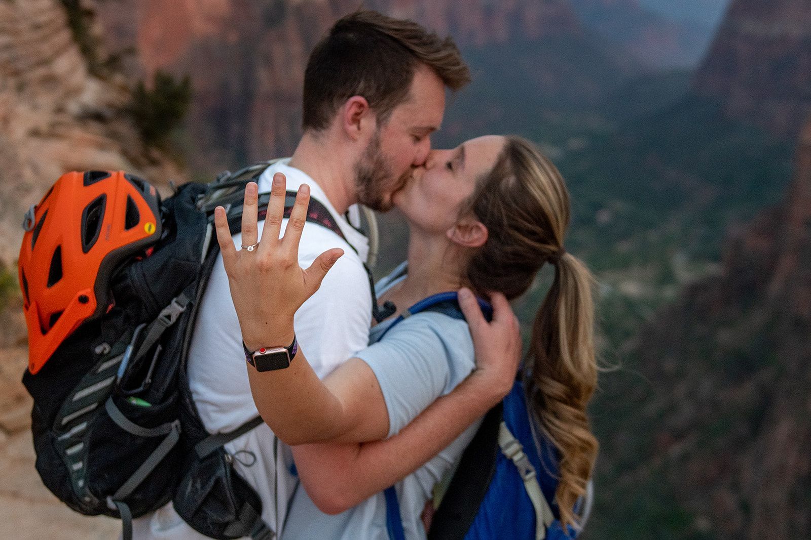 Two people kissing in a canyon and woman is folder up a finger with engagement ring on it