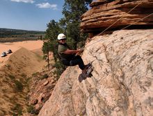Woman rappelling down a sandstone cliff, wearing a helmet and harness. Red desert landscape in background.