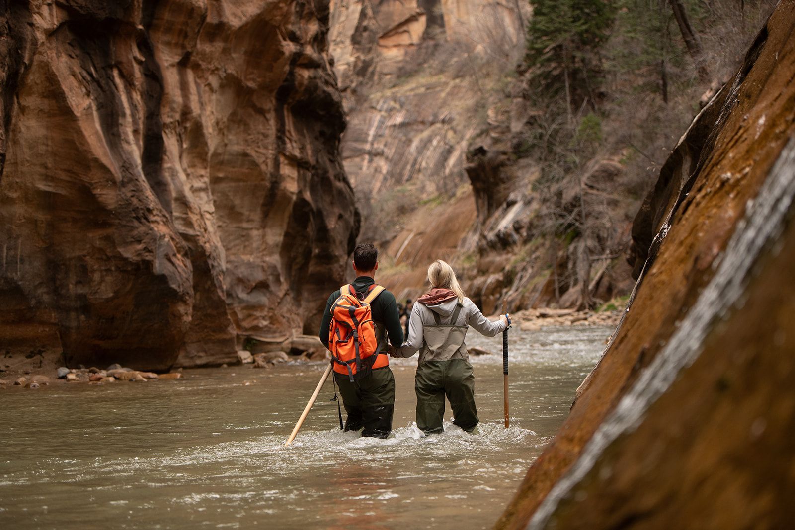 Two hikers wade through a shallow canyon river, one in an orange jacket and one using a walking stick.