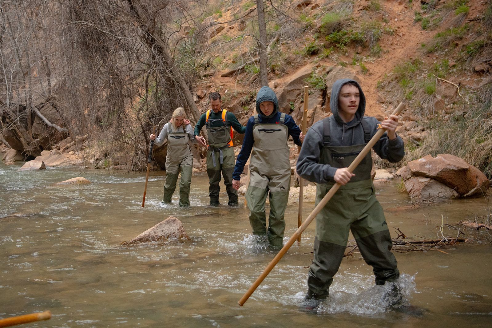 People wearing waders walk in a river with hiking poles