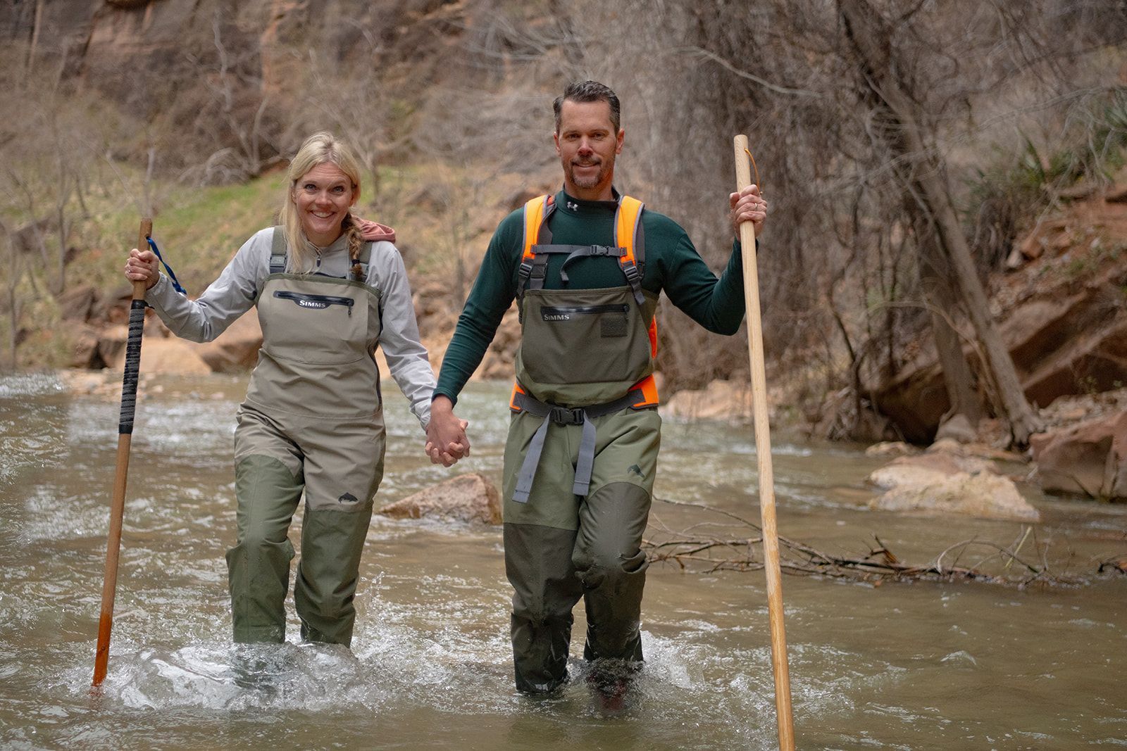 Two people holding hands walk through a river with towering cliffs lining the way