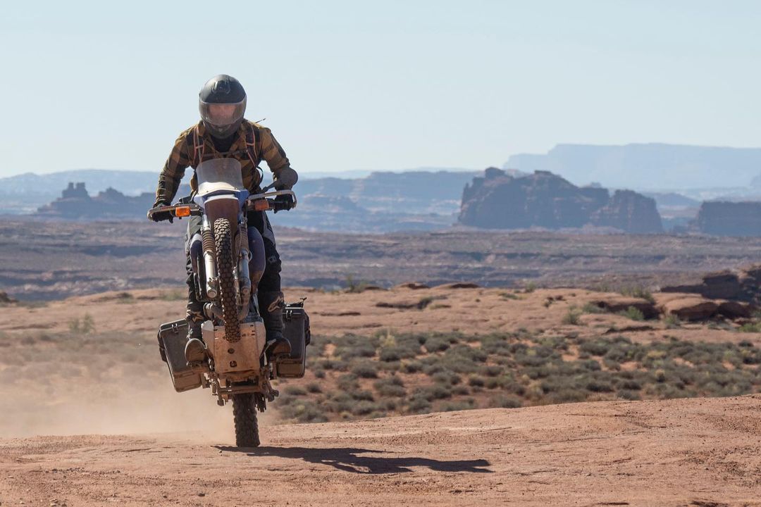 A man riding a dirt bike is doing a wheelie with rock formations in the background. 