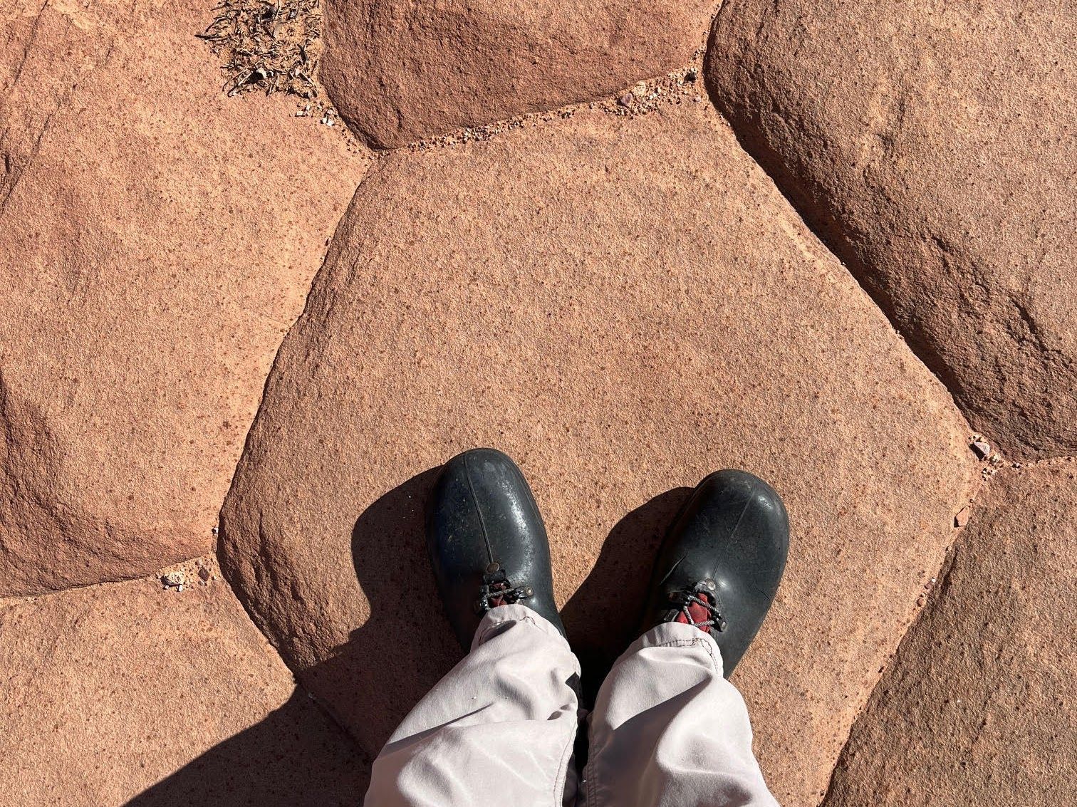 Person's feet in dark shoes standing on reddish-brown stone pavement.