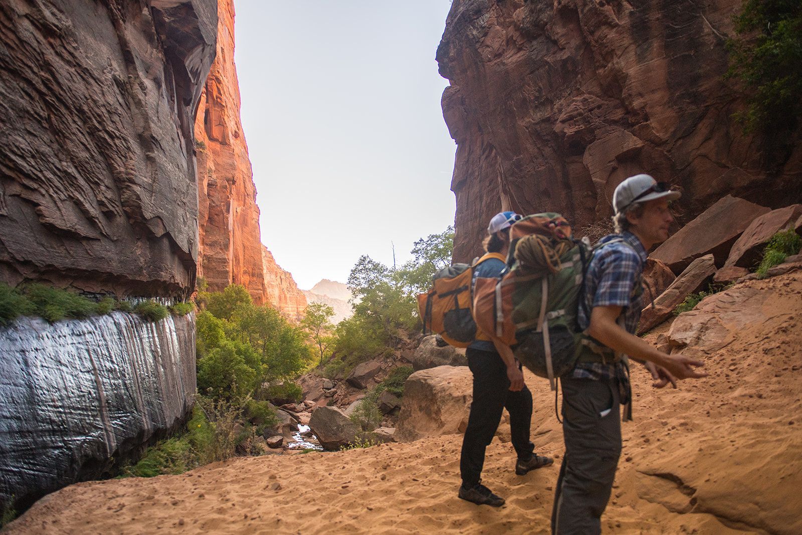 Hikers with backpacks walk through a narrow canyon, red rocks on either side, sunny sky above.