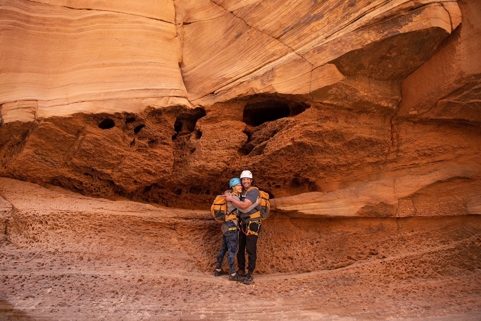 Couple in harnesses hugging in a sandstone canyon, lit by natural light.