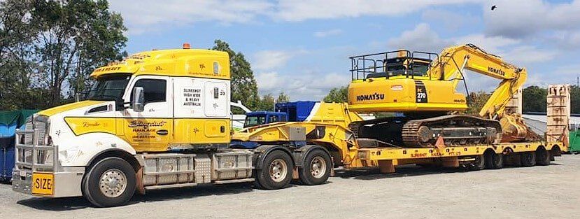 A yellow and white semi truck is carrying a large excavator on a trailer- Slingshot Haulage in Katherine, NT