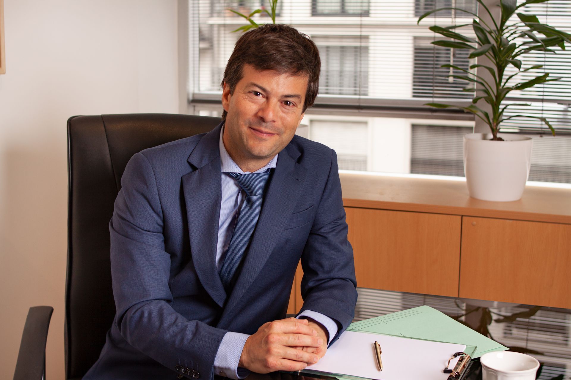 Un hombre con traje y corbata está sentado en un escritorio frente a una ventana.