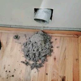 Lint pile on wooden floor under a vent opening in a ceiling.