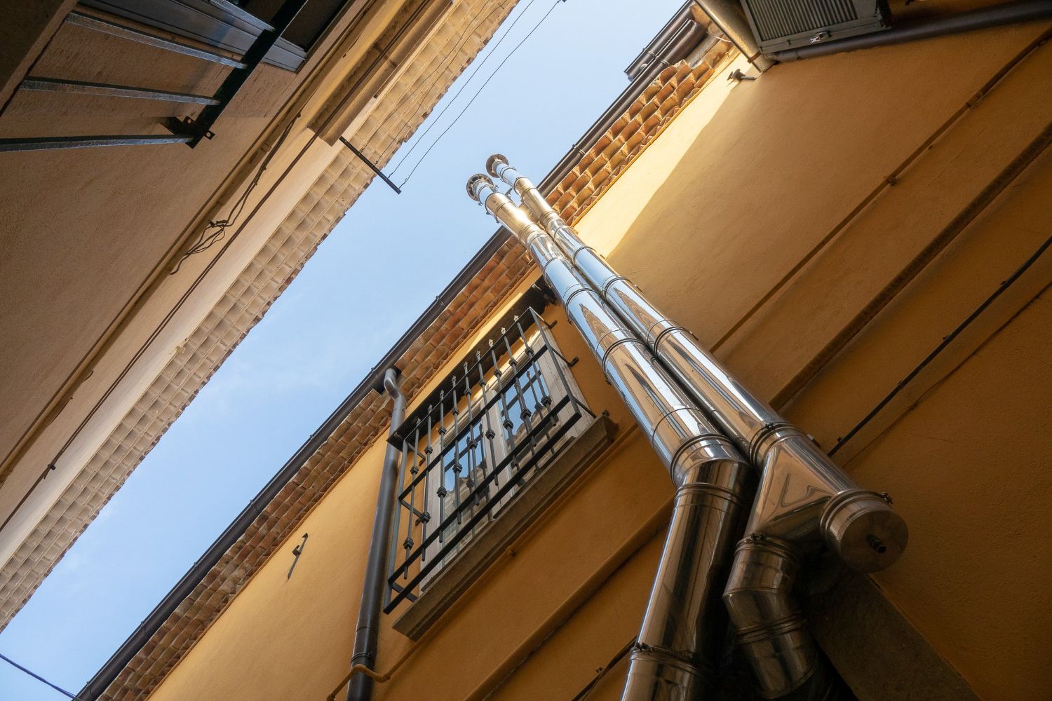 Upward view of a narrow alley with tan buildings, blue sky, and metal pipes