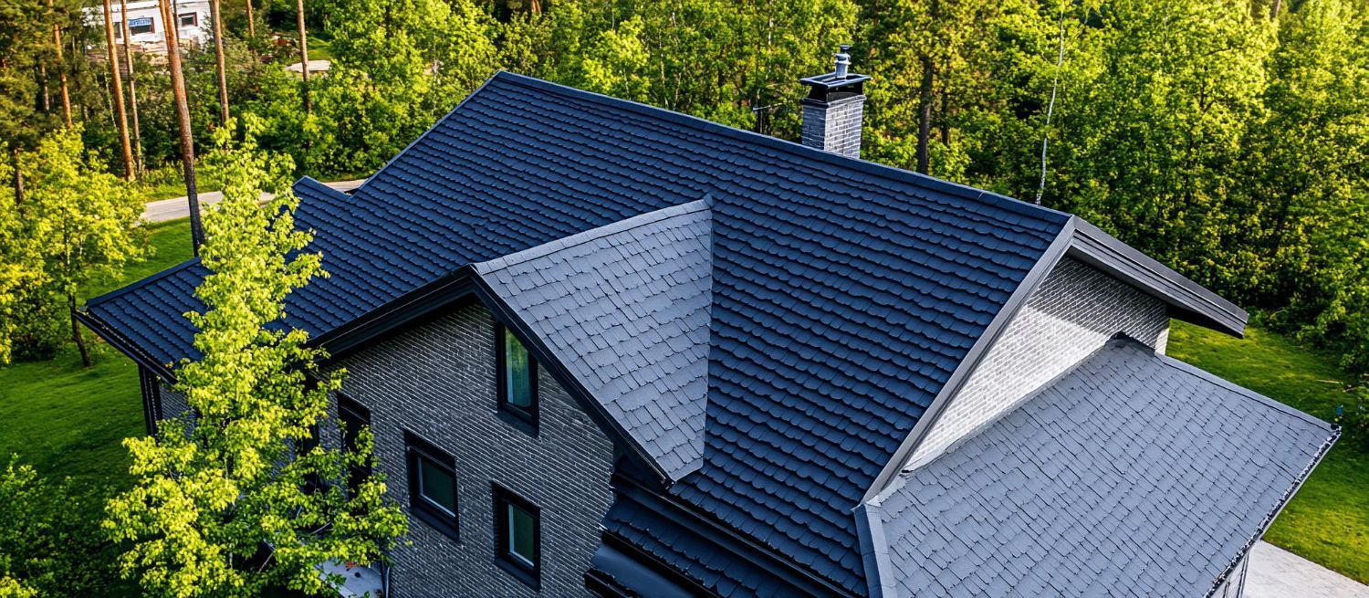 Aerial view of a gray house with a dark shingled roof surrounded by dense green trees.