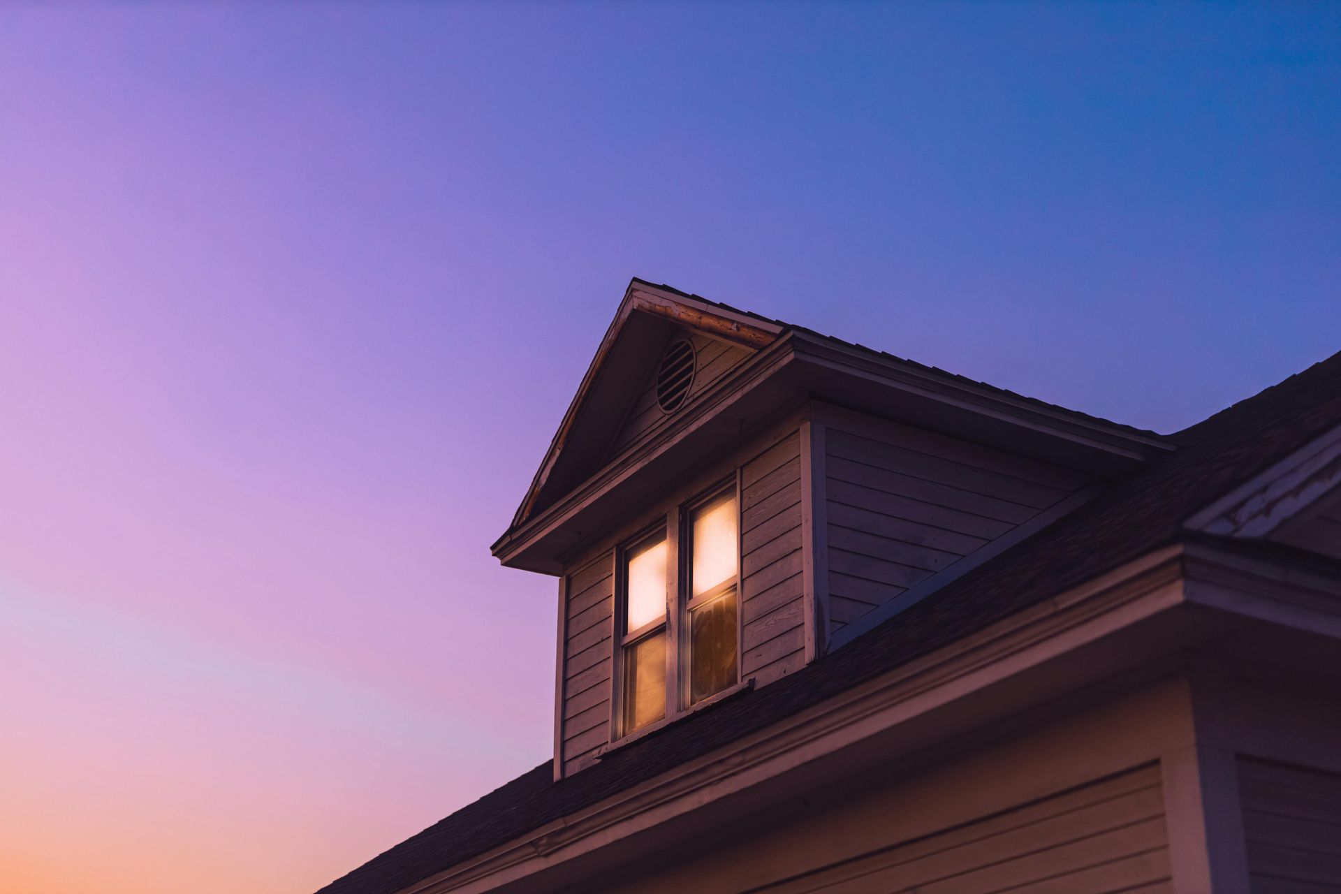 House roof at dusk with a glowing window against a purple-pink sky