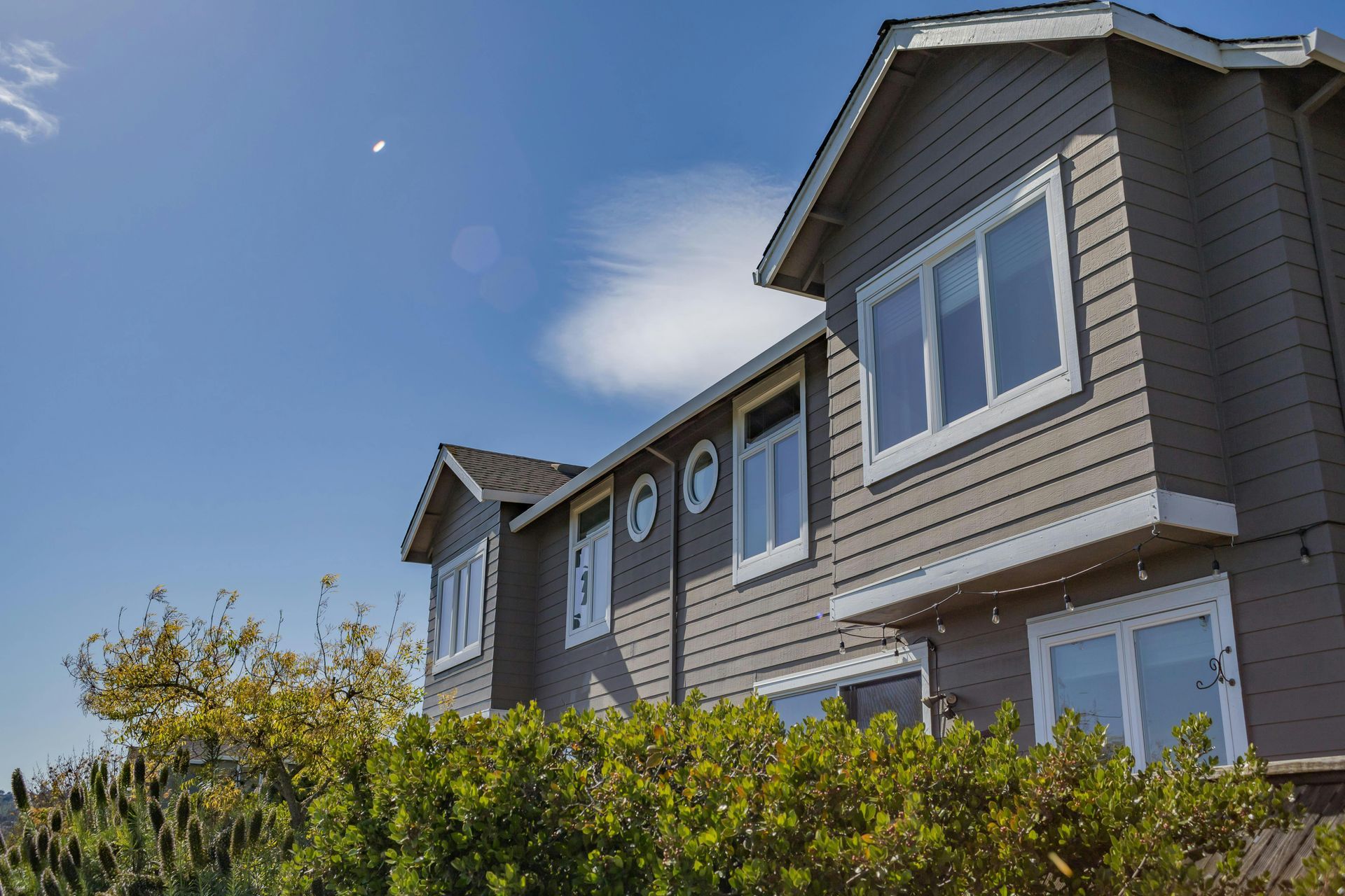 Modern gray house with white trim behind green bushes under a clear blue sky